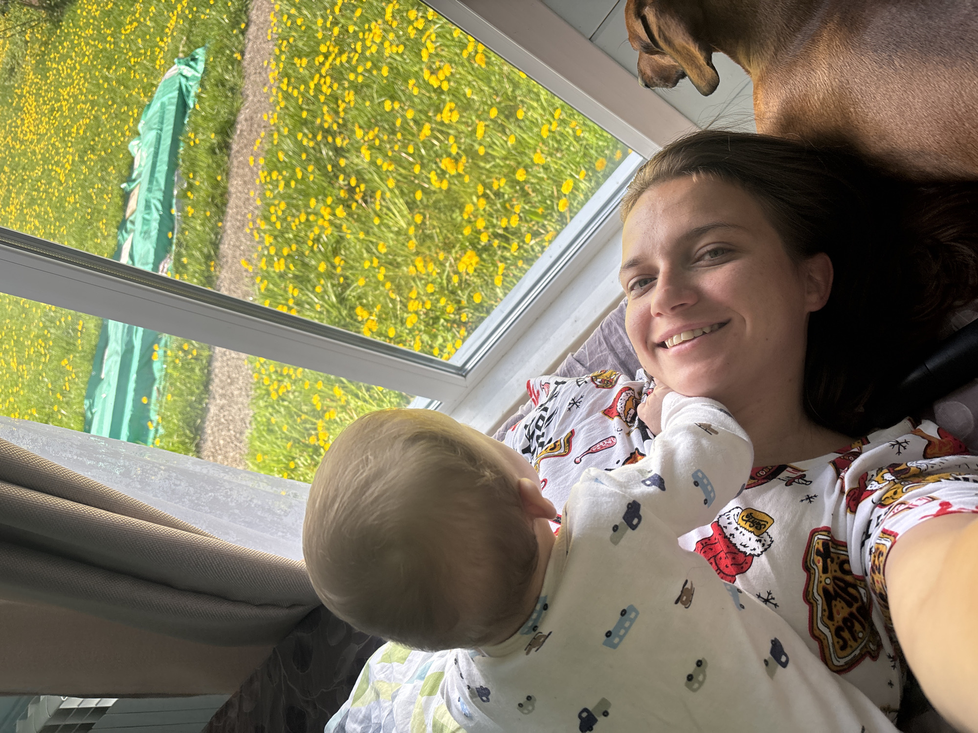 The photograph shows a woman taking a selfie while lying down with a baby. A brown dog is near the woman's head. Outside the window, there is a grassy field with yellow flowers, a sidewalk, and a green tarp. The woman is wearing pajamas and smiling. The baby is wearing a white onesie. The location is identified as Раздолье, Russia.