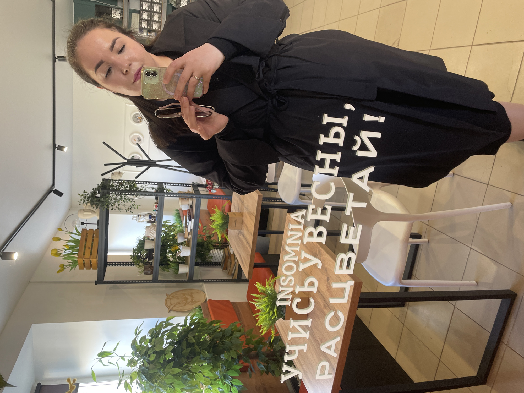 The image is a selfie of a woman in a black dress taking a picture of herself in a cafe, likely in the city of Chaikovsky, Russia, based on the Cyrillic text "INSOMNIA УЧИСЬ У ВЕСНЫ, РАСЦВЕТАЙ!" (INSOMNIA LEARN FROM SPRING, BLOOM!). The woman is holding a phone in her right hand and eyeglasses in her left hand. The background of the image features a cafe interior with wooden tables and white chairs. There is a shelving unit with plants and decorative items. The cafe has a light-colored floor and white walls. The lighting is bright, possibly natural light supplemented by artificial light from ceiling fixtures.