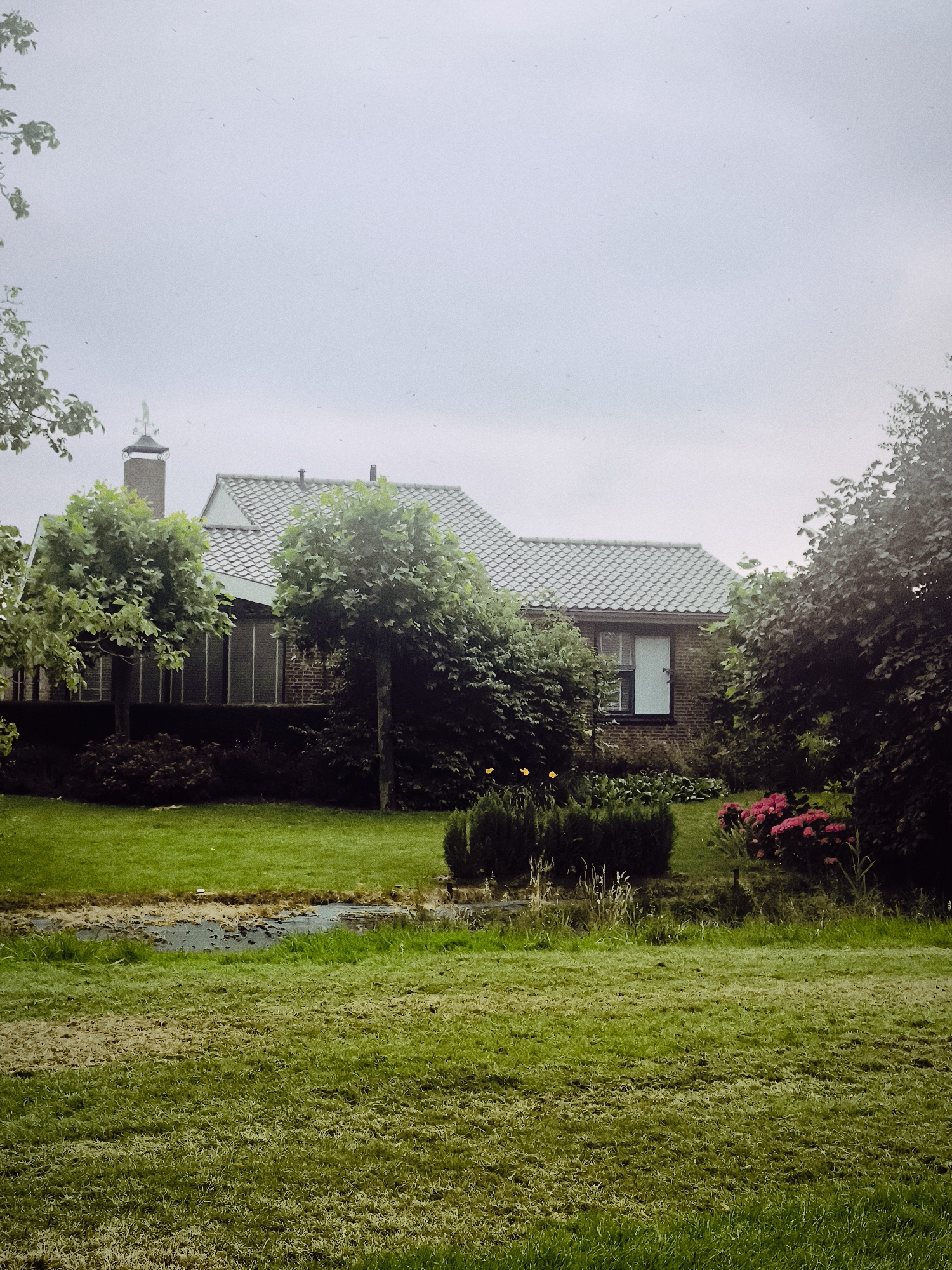 The image depicts a residential property in Almere, Netherlands, characterized by a brick house with a tiled roof. The house features a prominent chimney and large windows, suggesting a modern architectural style. Surrounding the house is a lush green lawn, meticulously maintained, with a small pond or water feature in the foreground. Dense foliage and trees frame the property, creating a sense of privacy and natural beauty. The sky is overcast, indicating a cloudy or possibly rainy day. The overall scene conveys a peaceful and idyllic atmosphere, typical of suburban or rural settings.