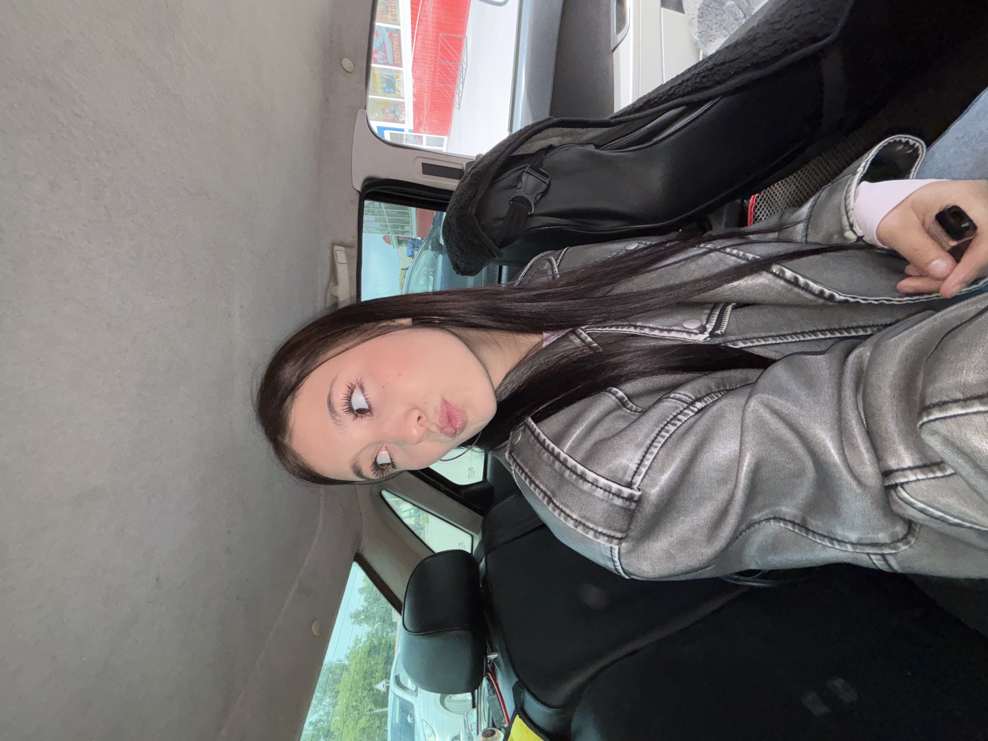 A young woman with long, dark brown hair and light-colored eyes is captured in a selfie taken inside a car, seemingly from the back seat. She is making a playful expression with puckered lips, looking upwards and to the side. She wears a silver-grey faux leather jacket with prominent stitching, a light pink or white long-sleeved top underneath, and light blue jeans. A black smartwatch or similar device is visible on her right wrist.
The car's interior features a grey headliner and black seating. A black bag with a visible buckle rests on the seat next to her, and part of a red seatbelt buckle is also visible. Through the car windows, an outdoor scene unfolds, revealing other vehicles, including a white van or car, and some green foliage from trees. In the background, a building with distinct red elements is visible. Faint Russian text, "АВТО АВТОБ", likely referring to "Auto" or "Automobile," can be seen on a sign on this building, indicating a commercial area.
The scene takes place during the daytime, with natural light illuminating the interior. The woman's pose and expression suggest a casual, lighthearted, or playful mood. The image provides a glimpse of a moment during travel or while parked in Chaikovsky, Russia.