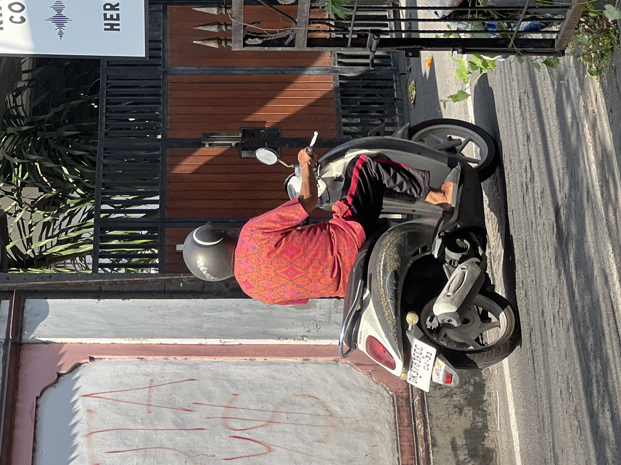 In Kuta, Indonesia, a person wearing a red patterned shirt, black pants with a red stripe, and a black helmet rides a scooter on an asphalt road. The scooter is a light gray and black model with a visible license plate that reads "DK3781FCC 04-31". To the left of the scooter, a textured, pink and white wall is visible with red graffiti. Behind the scooter, a dark metal gate with wooden planks and greenery is present. A white sign with the text "CO", a waveform graphic, and "HER" is also visible. The sun casts strong shadows across the scene.