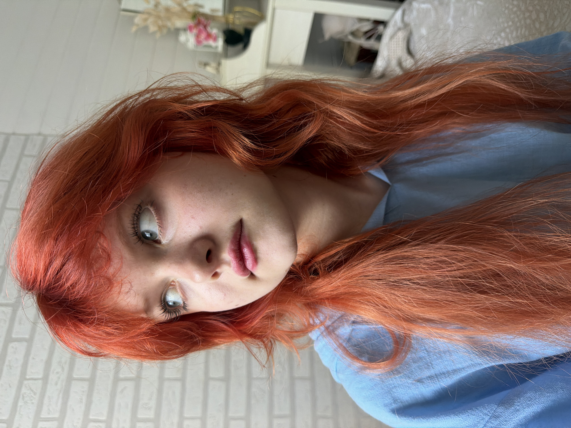The media file is a portrait of a young person with bright red hair. The person is looking upwards and to the left of the frame, with their mouth slightly open. Their eyes are light-colored and they appear to be wearing mascara. Their lips are a soft pink color. The background is out of focus, but appears to be an indoor setting with white brick-like walls and some decorative items on a shelf. The person is wearing a light blue garment. The lighting suggests it is daytime. The overall mood is relaxed and intimate. The image is oriented sideways. There is no visible text.