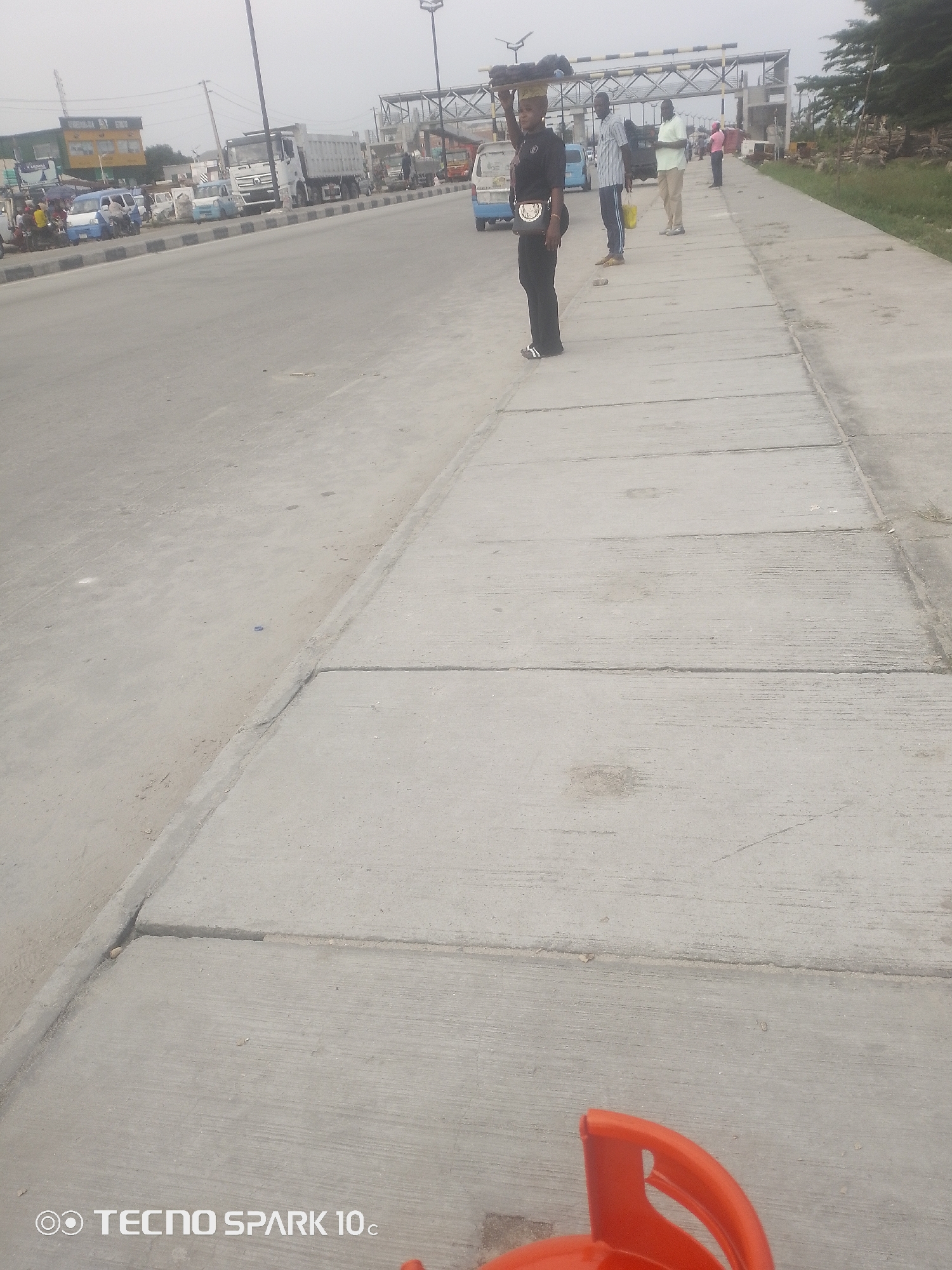 The image shows a street scene in Lekki, Nigeria, likely during the daytime, with an overcast sky suggesting cloudy weather. The foreground features a concrete sidewalk with visible expansion joints, and a portion of an orange gas cylinder or similar object is visible at the bottom right.
Several people are present on the sidewalk. A woman in the mid-left of the image is carrying a load of goods on her head, walking away from the camera. She is wearing dark clothing and a crossbody bag. Further down the sidewalk, a group of men are standing, seemingly waiting or observing. One man in a striped shirt carries a yellow bag. Another man in a light-colored shirt stands with his hands in his pockets. A third man in a light pink shirt is further down the sidewalk, also standing.
Behind the people, a road with traffic is visible. A large white truck is parked on the road, and other vehicles, including a blue minivan, are present. In the background, there is a structure that appears to be a toll booth or a bridge under construction, indicated by metal beams and scaffolding. Beyond the road and structures, there is some greenery and trees.
The overall impression is a busy, functional roadside environment in a Nigerian urban or semi-urban setting, with people engaged in daily activities. The presence of vendors and the general atmosphere suggest a typical day. Visible text includes "TECNO SPARK 10c" at the bottom left of the image, indicating it's a phone's watermark.