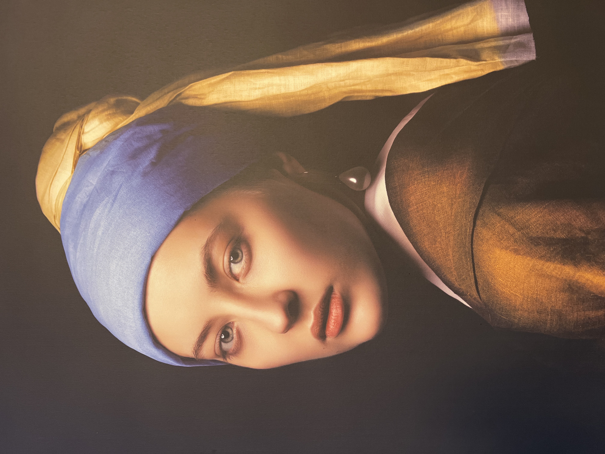 The image is a close-up, posed portrait of a young woman with light skin and striking blue eyes. She is wearing a blue headscarf that covers her hair, with a yellow fabric draped over it and hanging down her side. She also wears a dark brown garment with a white collar and a large, teardrop-shaped pearl earring in her left ear. Her gaze is directed to the viewer's left. The background is dark and featureless, with the lighting focused on the woman's face and the textures of her clothing and headwear. There is no discernible information in the image to indicate a location in the city of Vitsebsk, Belarus.