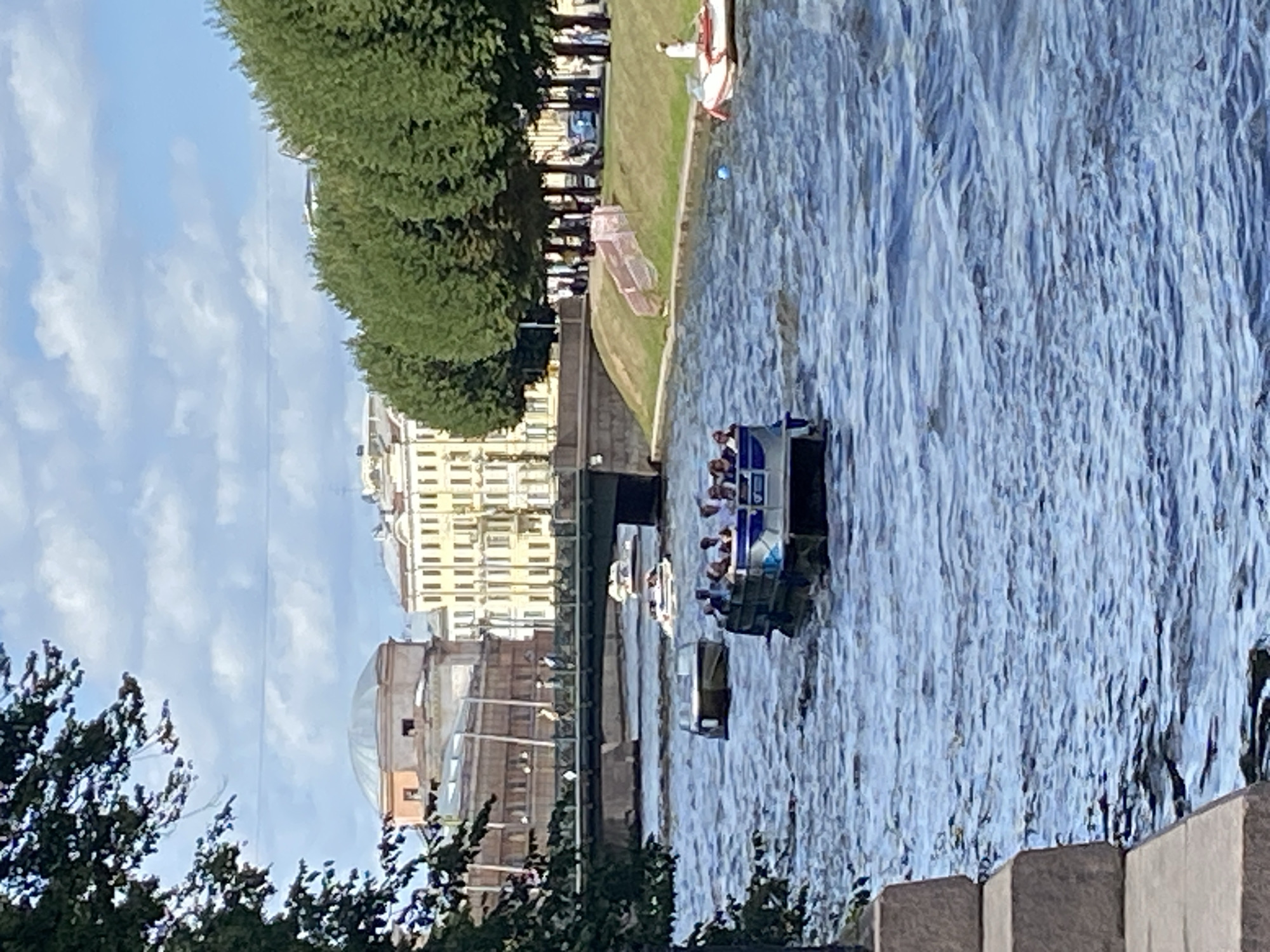 A wide shot captures a sunny day with puffy white clouds against a pale blue sky over a river in Saint Petersburg, Russia. A leafy green tree branch hangs into the frame from the upper left corner. The river is a choppy, deep blue, with whitecaps and reflections. In the middle ground, several boats navigate the water. The largest, a tour boat filled with people, is in the foreground, its dark hull and white upper deck clearly visible. Behind it, a smaller, lighter-colored boat heads in the opposite direction. Further in the distance, a bridge spans the river, with people walking across it. On the far bank of the river, a line of lush green trees separates the water from a row of classical, cream-colored buildings, some with distinctive rounded roofs and dormer windows. The architecture suggests a historic part of the city, likely along one of its many canals or rivers.