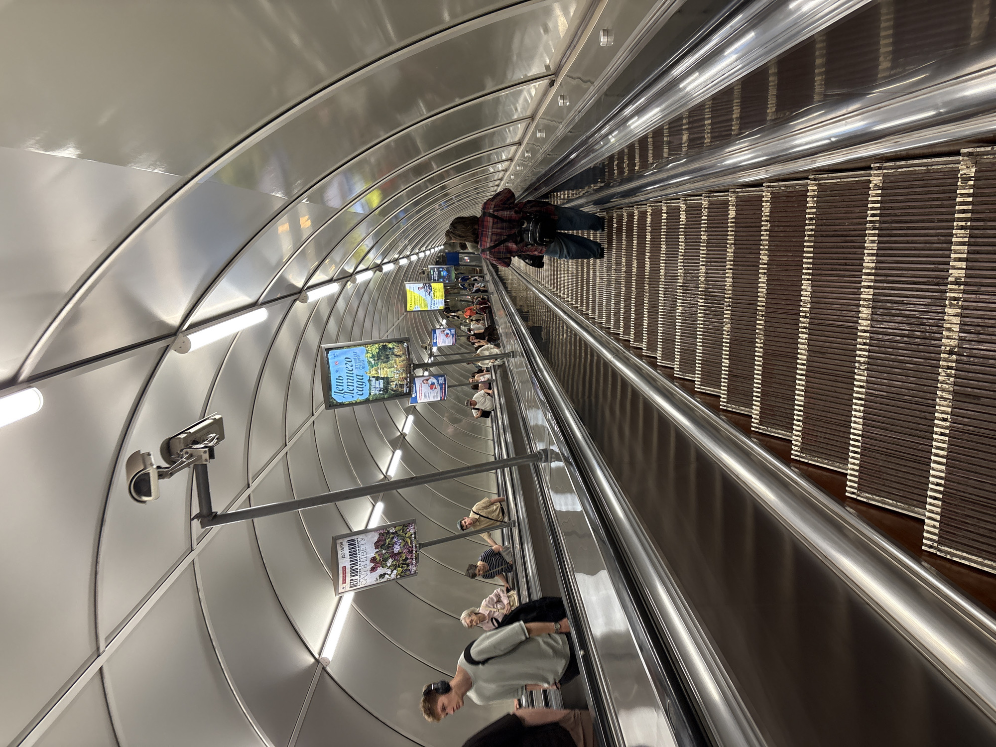 The image shows the inside of an escalator tunnel. The tunnel has a curved, metallic ceiling with evenly spaced lights. People are riding the escalator in both directions. Advertisements are mounted along the ceiling. A security camera is visible on the left side of the frame. The environment is enclosed and brightly lit.