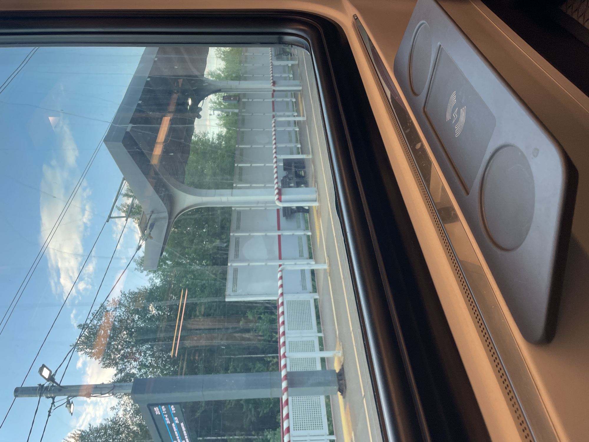 The image, rotated 90 degrees clockwise, captures a view from inside a modern train, looking out a window at a station platform.
**Subjects:**
- **Inside the train:** A thick black window frame occupies the left side of the image. To its right, a light-colored interior panel houses a grey wireless charging pad, marked with a white lightning bolt symbol and two circular indentations. A metallic, perforated strip, likely a vent or speaker grill, runs along the window's base.
- **Outside the train:** Through the window, a train station platform is visible. A red and white striped pole stands vertically on the platform, bordered by white panels or fences. A large, modern, light-grey canopy or roof structure extends over the platform. Further left, lush green trees are seen under a clear blue sky with scattered white clouds. A utility pole with lights and power lines crosses the lower left section. A digital information display board is partially visible at the bottom left.
**Activity or event:** The train appears to be stationary at a station platform. No people are visible.
**Notable details:**
- **Time of day:** Daytime, indicated by the bright sky and clear lighting.
- **Weather:** Clear and sunny with scattered clouds.
- **Location cues:** The modern design of the train interior and station structure suggests contemporary public transport. The text on the display board specifies destinations in Russia, consistent with the provided location context of Gribanovo, Russia.
**Visible text:**
- On the charging pad: A stylized white lightning bolt, indicating wireless charging.
- On the partially visible digital information display board (bottom left, rotated): "MOSKVA" (Moscow), "BALTIYSKAYA", "D_P-211-1", "1905", and fragments such as "OTPA" (likely part of "Отправление" - Departure) and "PRIP" (likely part of "Прибытие" - Arrival). Several times such as "20:00", "20:05", "20:10", and "20:20" are also visible.