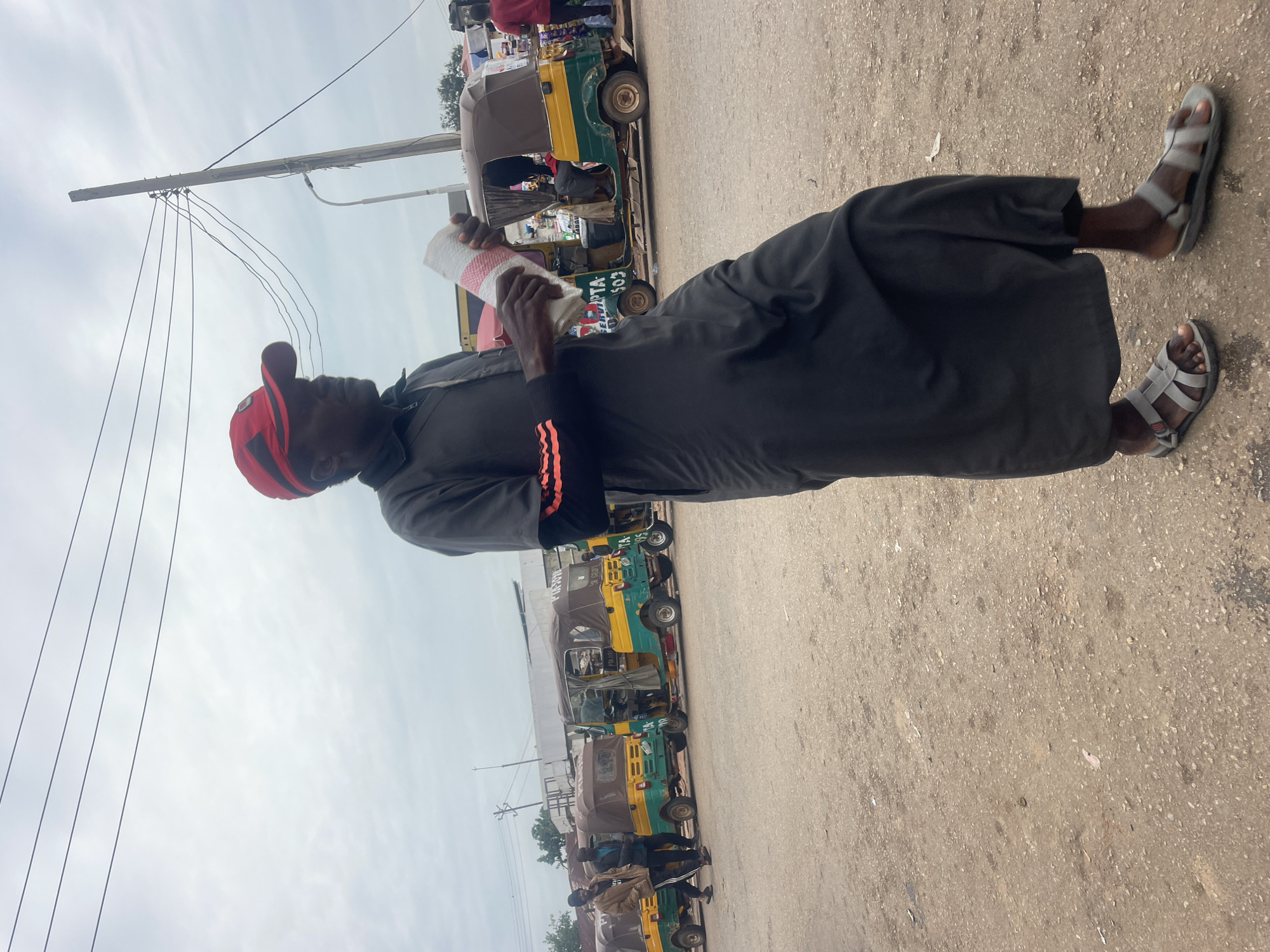 A medium shot captures a man standing in what appears to be a busy transportation hub or marketplace in Gura Top, Nigeria. The man, wearing a dark gray tunic-like garment, dark pants, gray sandals, and a red baseball cap, holds a rolled-up white and pink object in his right hand. His left arm is bent, and his hand is visible near his chest. He is looking to his right, away from the camera.
Behind him, a row of yellow and green auto-rickshaws, a common mode of transport in the region, are parked. Some of the rickshaws have canopies and appear to be waiting for passengers. Several other individuals are visible in the background, some standing and others near or inside the rickshaws. The ground is a mixture of dirt and gravel, suggesting an unpaved area. Electrical wires are strung across the sky, supported by wooden poles, indicating urban infrastructure. The sky is overcast with clouds. The overall scene suggests a typical day in a bustling Nigerian city.