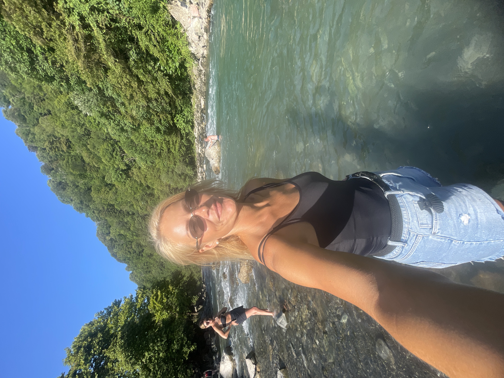 A blonde woman in a black tank top and denim shorts is taking a selfie by a river. She is wearing sunglasses and a pearl earring. Behind her, a second woman in a black bikini and skirt stands in the shallow water. Further in the background, a man in swim trunks is in the river. The river flows through a lush green mountainous landscape under a clear blue sky. This scene is consistent with the Mzymta River in Sochi, Russia, a popular destination for its natural beauty and recreational activities.