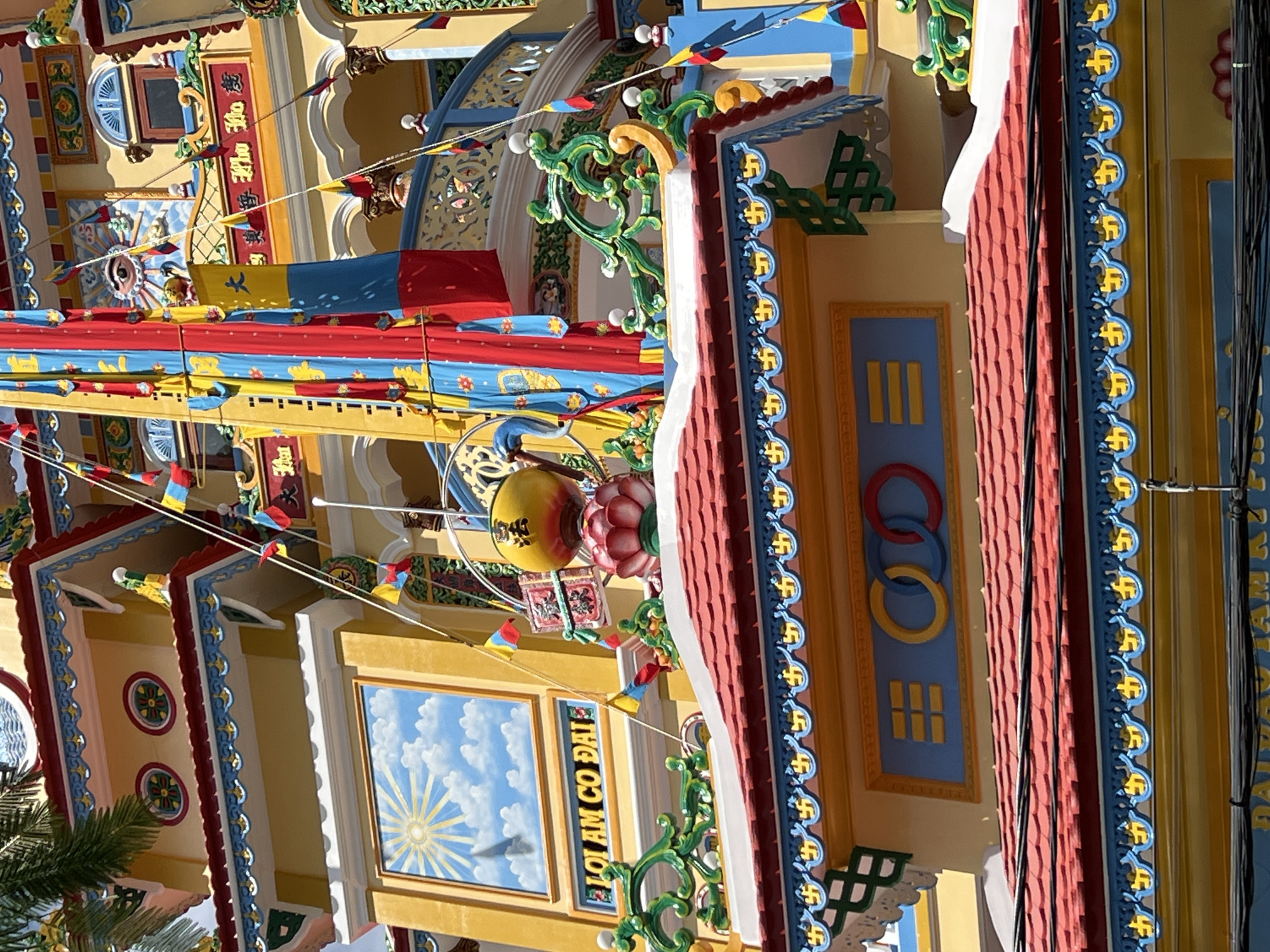 This is a detailed, forensic, matter-of-fact description of the provided image, which depicts a section of the Cao Dai temple in Nha Trang, Vietnam. **Scene:** A close-up view of the upper facade of a vibrantly colored building, likely a religious structure, under a bright sky. The architectural style is ornate and distinctly Asian, characterized by rich yellow walls, red tiled roofs, and intricate blue and yellow decorative trim. **Architectural Details:** * **Walls:** Predominantly painted a bright, sunny yellow, with accents of red and blue. * **Roofs:** Steeply pitched and covered with overlapping red tiles. A prominent decorative frieze runs along the eaves, featuring a repeating blue and yellow pattern that appears to be stylized swastikas, a common symbol in many Asian religions. * **Decorative Elements:** The building is adorned with numerous decorative features, including: * Green, ornate, scroll-like sculptures resembling stylized foliage or dragon heads, placed on ledges. * Small flags or banners in red, yellow, and blue, strung on wires, adding to the festive or spiritual atmosphere. * A large, yellow, spherical object, possibly a lantern or ornament, with a red lotus flower-like base beneath it. This object is positioned on a higher level of the building. * A mural depicting a sun with rays and clouds, enclosed in a rectangular frame. Below this mural is a blue band with yellow characters, possibly a sign or inscription. * On a lower level, a blue rectangular panel features a motif of three interlocking rings (yellow, blue, and red) and a set of horizontal yellow bars. * Balconies or elevated walkways with blue railings and ornate white and green supports are visible on upper levels. * Round windows with patterned interiors and arched windows with decorative grilles are present. **Symbolism and Potential Interpretations:** * The presence of swastikas, the lotus flower, the sun motif, and the vibrant colors are indicative of a Buddhist or syncretic religious context, consistent with Cao Dai practices. * The interlocking rings could be a secular or spiritual symbol of unity or interconnectedness. * The flags suggest ceremonial or celebratory events. **Environment:** The image appears to be taken outdoors, with natural light illuminating the structure. The presence of a palm frond in the upper left corner indicates a tropical or subtropical climate, fitting for Nha Trang, Vietnam. No people are visible in the frame. The focus is solely on the architectural details and decorations of the building.