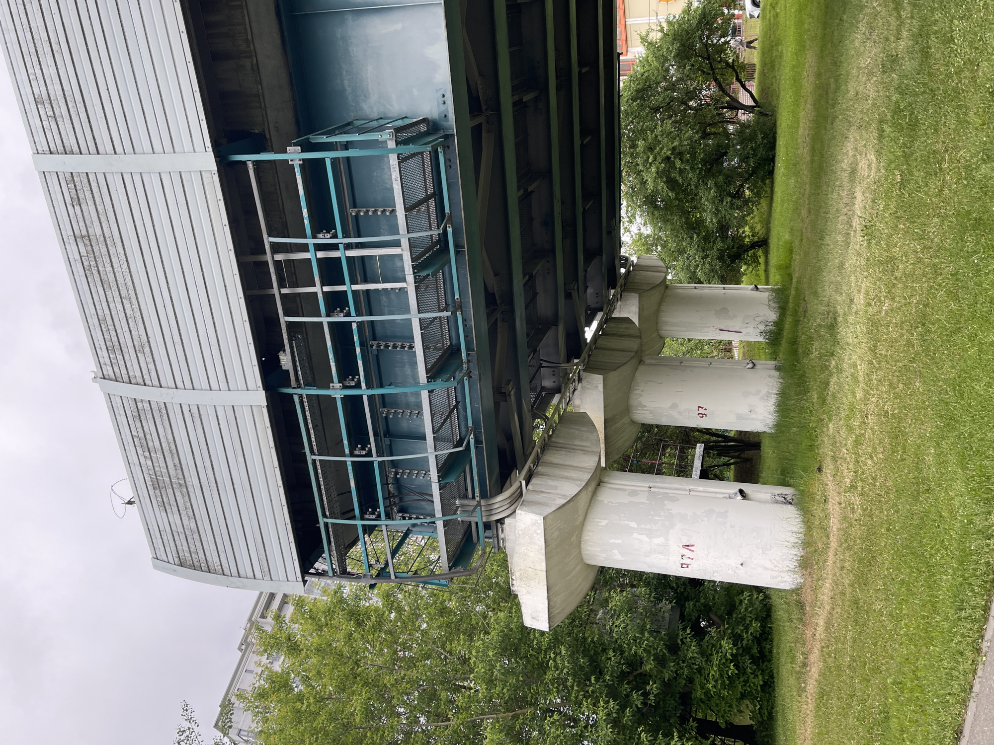 A low-angle shot depicts the underside of the Moscow monorail track, supported by white cylindrical pillars on a green lawn. The monorail structure features a gray corrugated roof and a blue metal framework, including a walkway. The pillars are marked with red numbers "97" and "97A." Trees are visible in the background along with parts of buildings. The sky is overcast.