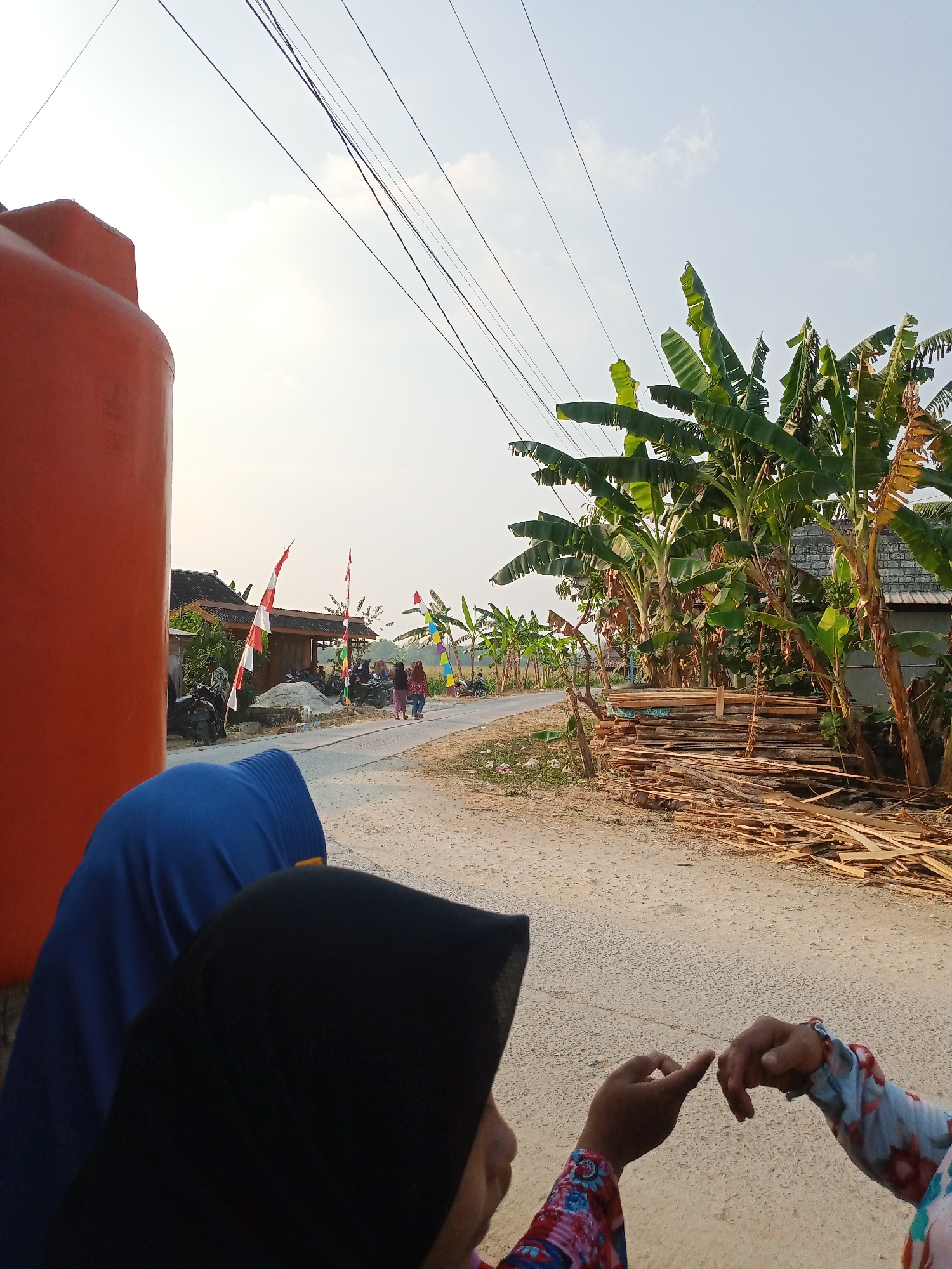 In Rowobungkul, Indonesia, an outdoor scene unfolds with a focus on a dusty road and its surroundings. In the foreground, two individuals are partially visible, their heads covered by blue and black hijabs. Their hands are interacting, one with a pointing finger and the other with an outstretched hand, suggesting a conversation or gesture. To the left, a large, bright orange water tank dominates the frame. The road stretches into the distance, lined with banana trees, some laden with fruit. A pile of cut wood rests beside the road. Further down the road, several motorcycles are parked, and small buildings with red and white flags are visible. The sky above is a pale blue with scattered clouds, and power lines crisscross overhead. The overall impression is one of a rural Indonesian setting with everyday elements of life.