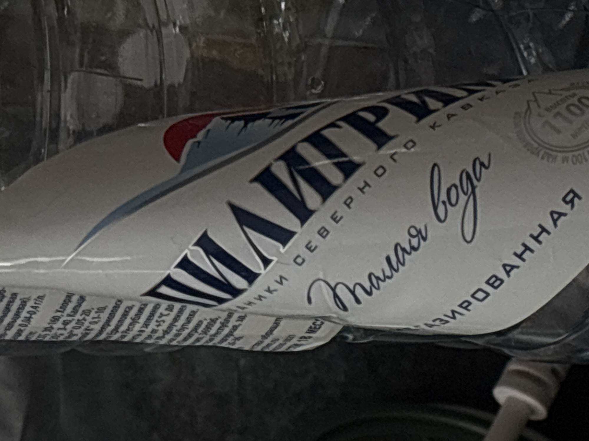 The image shows a close-up of a plastic bottle of mineral water. The label is predominantly white with dark blue text and some red and blue graphic elements. The text on the label is in Russian and indicates the brand name "Пилигрим" (Pilgrim), followed by "СЕВЕРНОГО КАВКАЗА" (Northern Caucasus), and then "Малая вода" (Small water). Below this, it says "ГАЗИРОВАННАЯ" (sparkling). There are also some nutritional or mineral content details visible on the left side of the label. The bottle appears to be partially filled with clear liquid. The background is dark and out of focus, suggesting the photograph was taken indoors in low light conditions. There are no people or discernible city landmarks visible in the image. The location in Saint Petersburg, Russia, is not identifiable from the provided image.