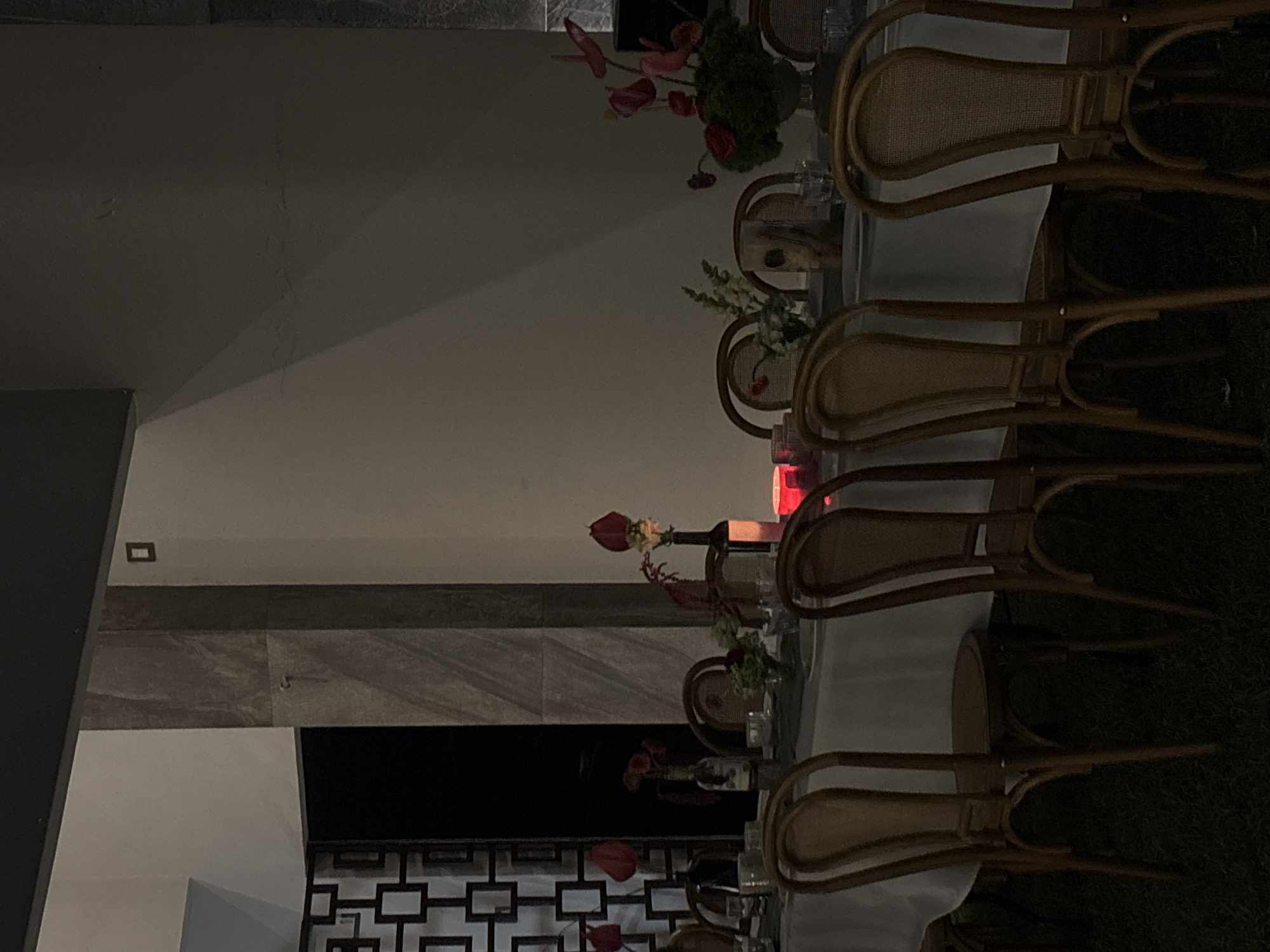 The image displays an unoccupied outdoor or semi-outdoor event setup at night. A long table draped with a white tablecloth and a dark grey runner is central to the scene. Numerous empty wooden chairs with cane backs are arranged along both sides of the table, resting on a dark, indistinct ground surface, possibly grass. The table is set with clear drinking glasses and white plates. Multiple floral arrangements, predominantly featuring red anthurium flowers, are distributed along the table, some utilizing dark bottles as vases. A single red-glowing light source, likely a candle in a holder, is positioned on the table. The background includes a light-colored wall to the right, partially obscured by shadows, and a tall, grey-tiled column in the mid-ground. To the left, a dark area with a decorative metal screen featuring a geometric pattern is visible. No individuals are present. The specific location within the city of Puebla, Mexico, cannot be determined from the visual evidence provided.