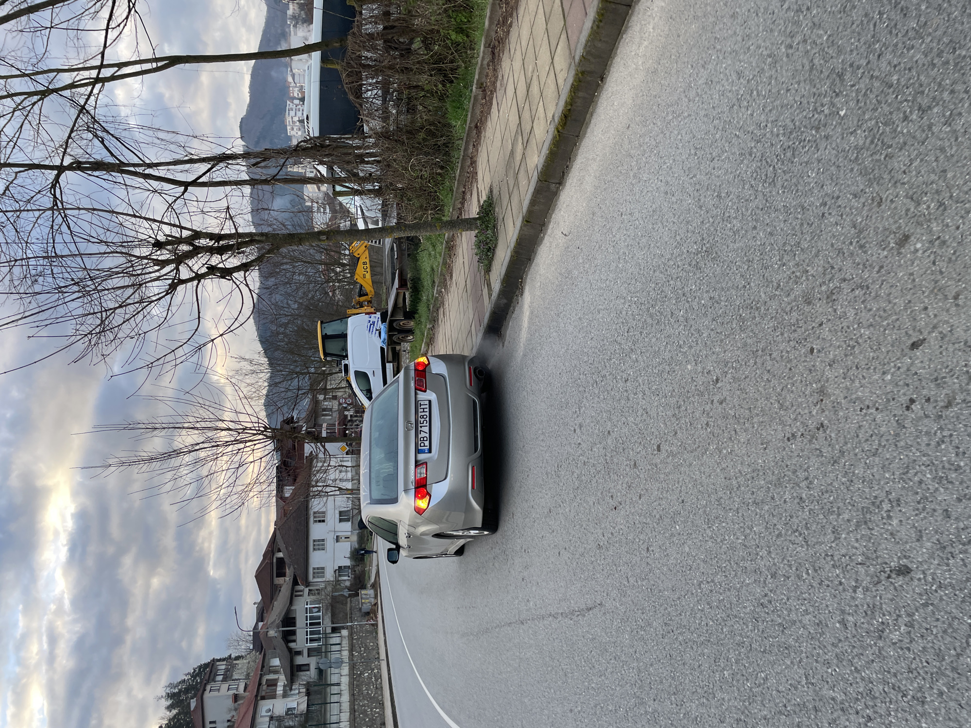 The image shows a silver Honda Accord with license plate "PB 7158 HT" parked on a road. The vehicle is partially obstructing the lane. A white van, potentially towing a small yellow construction vehicle (JCB), is parked further ahead on the sidewalk. The surrounding environment includes buildings, trees without leaves, a paved sidewalk, and a view of mountains in the background. The location appears to be a residential area on the outskirts of a town, based on the buildings and landscape features.