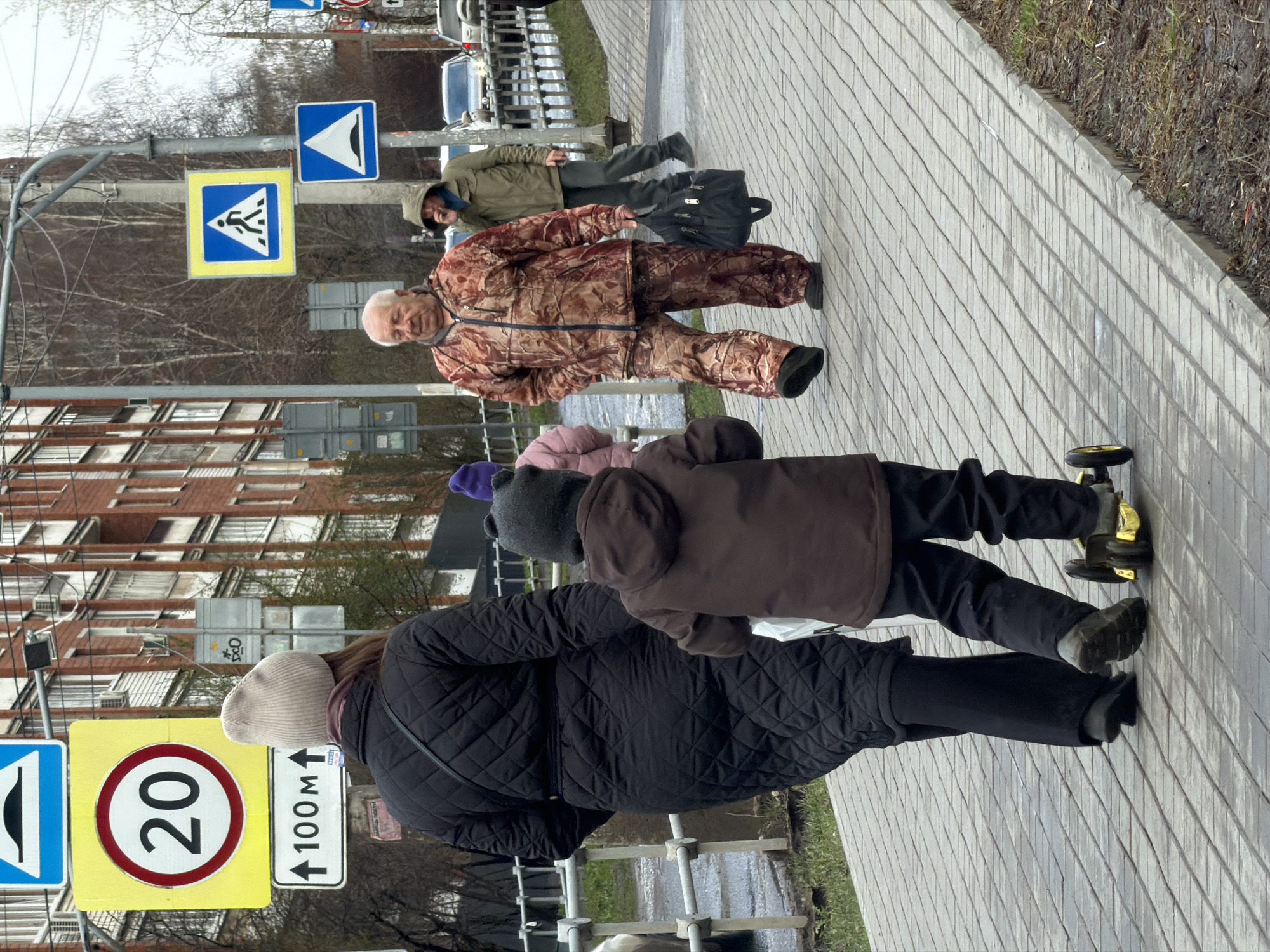 This image captures a street scene in Izhevsk, Russia, likely during an overcast day in a colder season given the bare trees and people's attire. The view is from a slightly elevated position, looking down a sidewalk that runs parallel to a road.
In the foreground, an adult, seen from behind, wears a black quilted jacket, dark pants, and a light-colored beanie. To their left, slightly ahead, a child in a brown jacket and dark beanie is seen from behind, pushing a small yellow and black ride-on toy with wheels. Further down the sidewalk, an older man with white hair and a mustache is walking towards the viewer, wearing a brown and green camouflage-patterned jacket and matching pants, with a neutral expression. Behind him, another person in a green hooded jacket and dark pants walks away from the viewer, carrying a black shoulder bag.
The sidewalk is paved with light grey, rectangular blocks. To the left of the sidewalk is a road, beyond which are bare-branched trees and multi-story, brick-colored apartment buildings. Several street signs are visible on poles alongside the road. These include a large yellow square sign with a red circular border containing the number "20", indicating a speed limit. Below it is a white rectangular sign with an upward arrow and the text "100M", likely denoting a distance or direction. Other signs include a blue square sign with a white humped road symbol, a yellow square sign with a blue border and a white pedestrian crossing symbol, and a blue square sign with a white downward-pointing arrow. The overall atmosphere is mundane and everyday.
