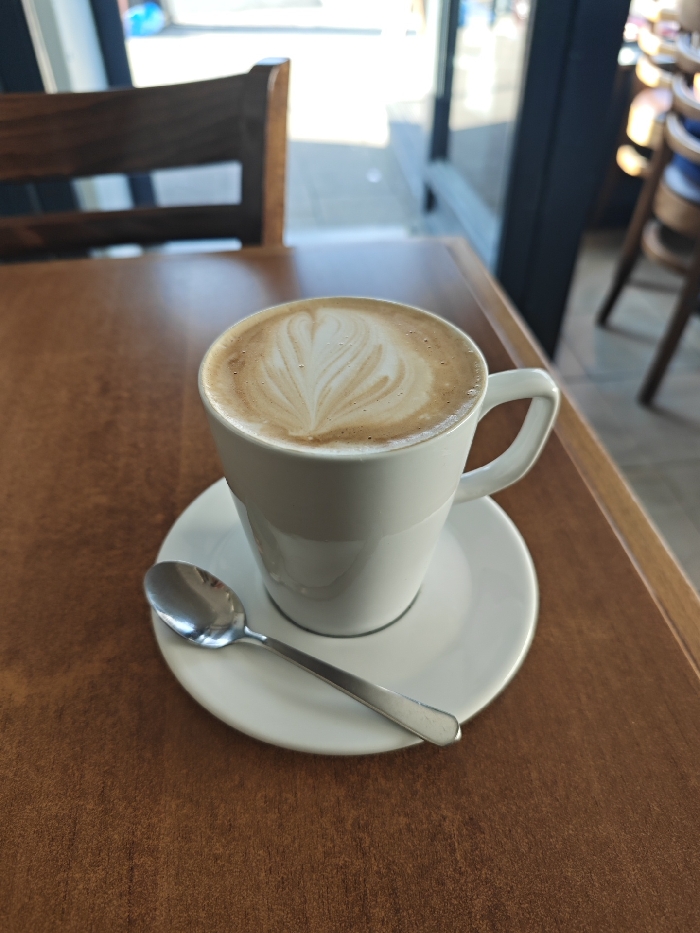 A white ceramic mug containing a latte with a prominent heart-shaped latte art design rests on a matching white ceramic saucer. A silver-colored spoon is positioned on the saucer beside the mug. These items are situated on a smooth, brown wooden table. The background indicates an indoor commercial establishment, specifically a cafe or restaurant, located in the City of London, United Kingdom. To the left, a dark brown wooden chair backrest is partially visible and out of focus. To the right, several other wooden chairs, some with indistinct blue elements, are present in the blurred background. A large window or glass panel allows bright natural light to illuminate the scene, offering a heavily blurred view of an outdoor urban environment beyond. No individuals are visible.