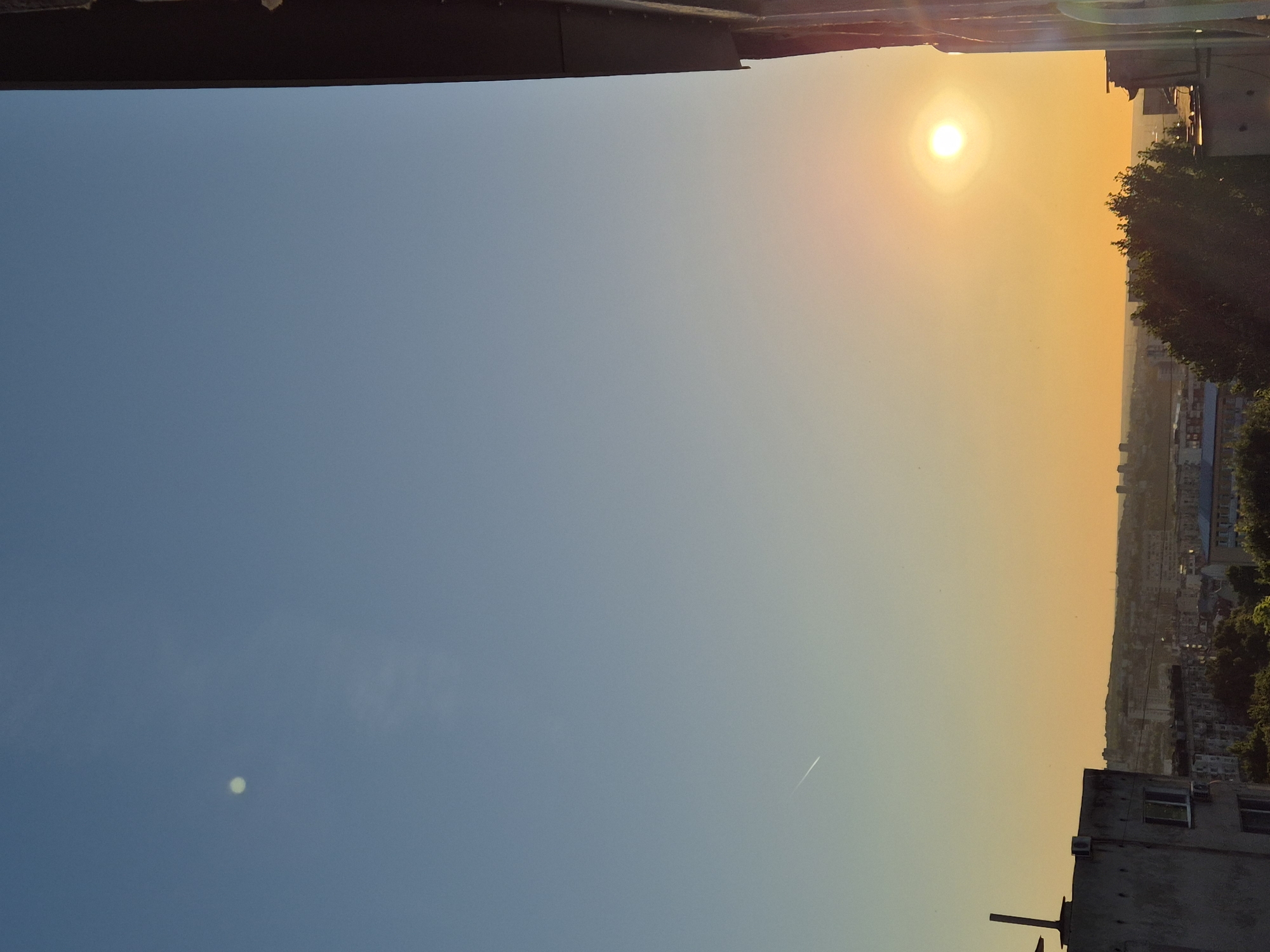The image depicts a city skyline at sunset, viewed from an elevated perspective. The sky transitions from a clear blue at the top to a warm orange hue near the horizon, indicative of the setting sun. The sun itself is a bright, distinct orb in the right-center of the frame. A faint white streak, possibly from an airplane, is visible in the upper left quadrant of the sky.
In the foreground, the dark silhouette of a building's edge frames the right side of the image. The middle ground features a densely packed urban landscape with numerous buildings of varying heights and architectural styles. Trees are scattered throughout the cityscape, with a cluster of darker foliage towards the bottom right. On the left side of the image, a portion of a concrete building with visible windows and air conditioning units is present. The overall scene is one of a typical urban environment bathed in the warm glow of dusk.