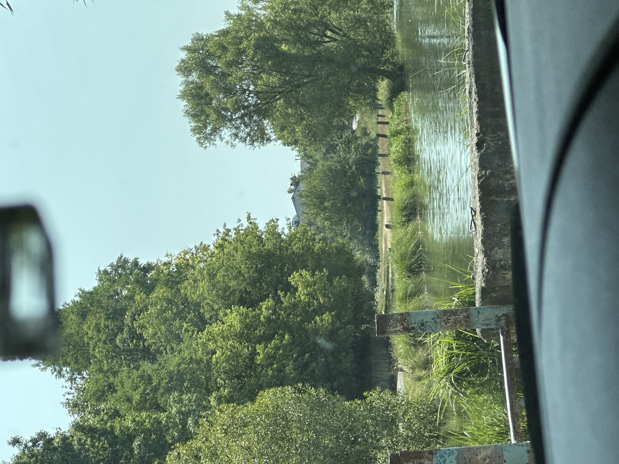 The image shows a body of water surrounded by lush green trees and vegetation. In the foreground, there is a concrete barrier with rusted metal posts. The water has a rippled surface, reflecting the clear blue sky. The background features a line of wooden posts along the bank, with a road visible behind them. Further in the distance, there are buildings and more trees. The overall impression is a tranquil, natural setting within or on the outskirts of the city of Makiv, Ukraine.