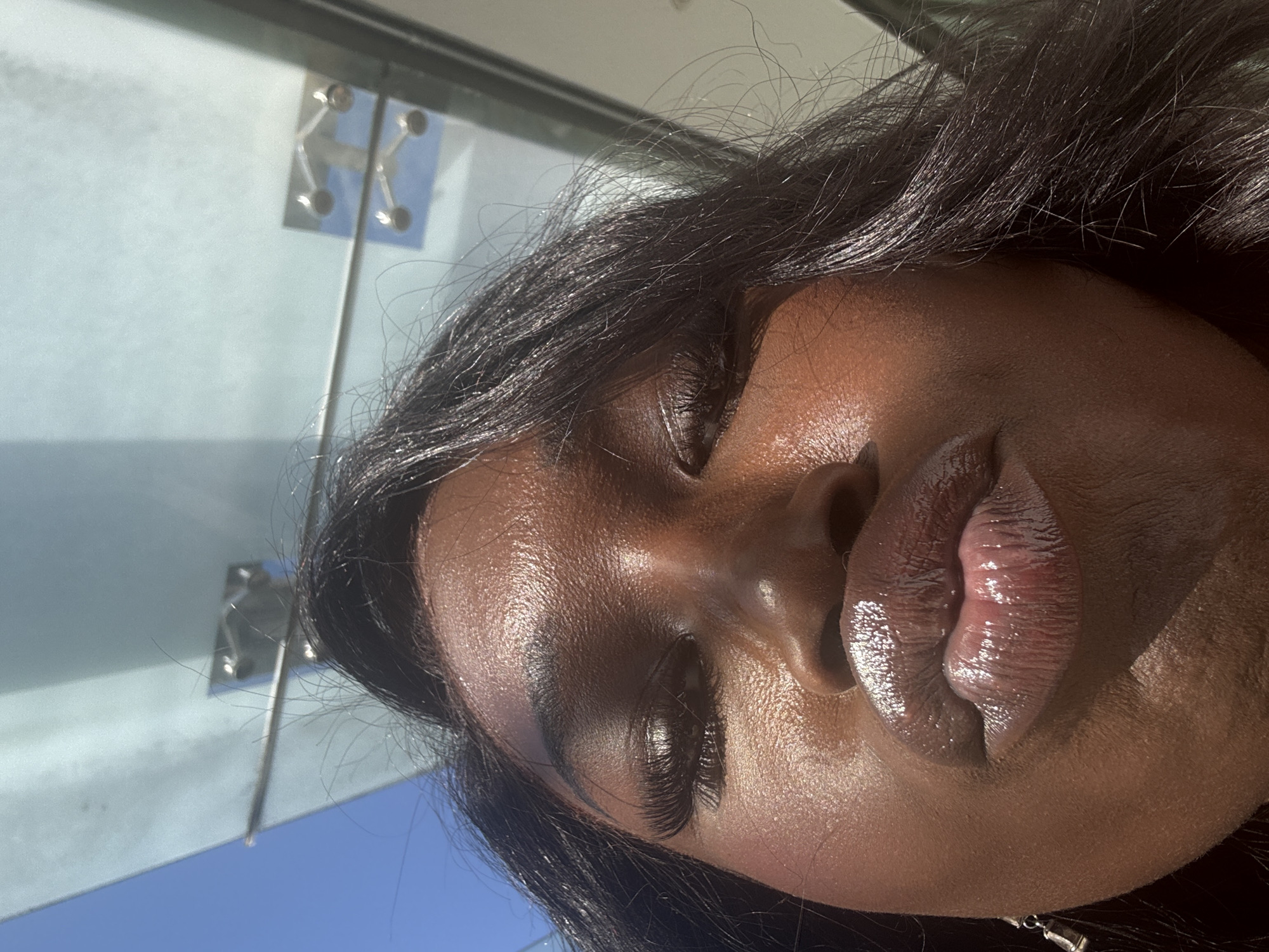 The image depicts a close-up of a Black woman's face, seemingly indoors, with part of a glass panel visible in the background. The glass panel is secured with metal fixtures. The woman's eyes are closed, and her hair is dark and styled. Her skin has a radiant sheen, and her lips are glossy and plump. The image's orientation suggests it may have been taken from a lower angle. The location is unknown without further information.