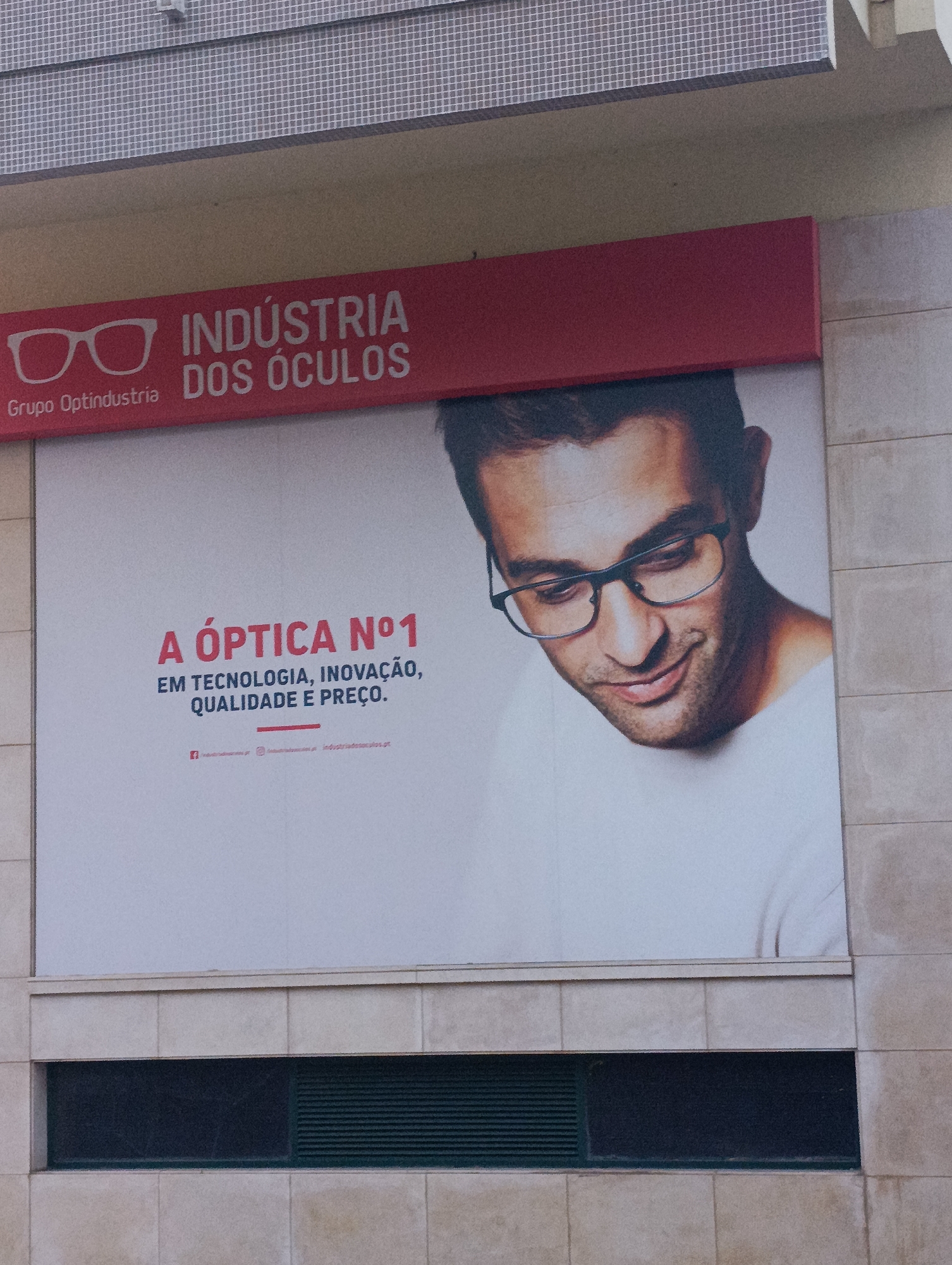 The image is a close-up, eye-level exterior shot of a billboard advertising "INDUSTRIA DOS ÓCULOS" (Eyeglass Industry) from "Grupo Optindustria". The billboard features a stylized graphic of eyeglasses and a portrait of a man wearing glasses, with the slogan "A ÓPTICA Nº1 EM TECNOLOGIA, INOVAÇÃO, QUALIDADE E PREÇO." (The #1 Optician in Technology, Innovation, Quality and Price.). Website and social media icons are visible at the bottom of the billboard. The billboard is mounted on a beige tiled building. The top of the image shows a section of the building's facade with a checkered pattern. The bottom of the image reveals a dark rectangular vent. This billboard is located on Avenida Manuel da Maia in Setúbal, Portugal.