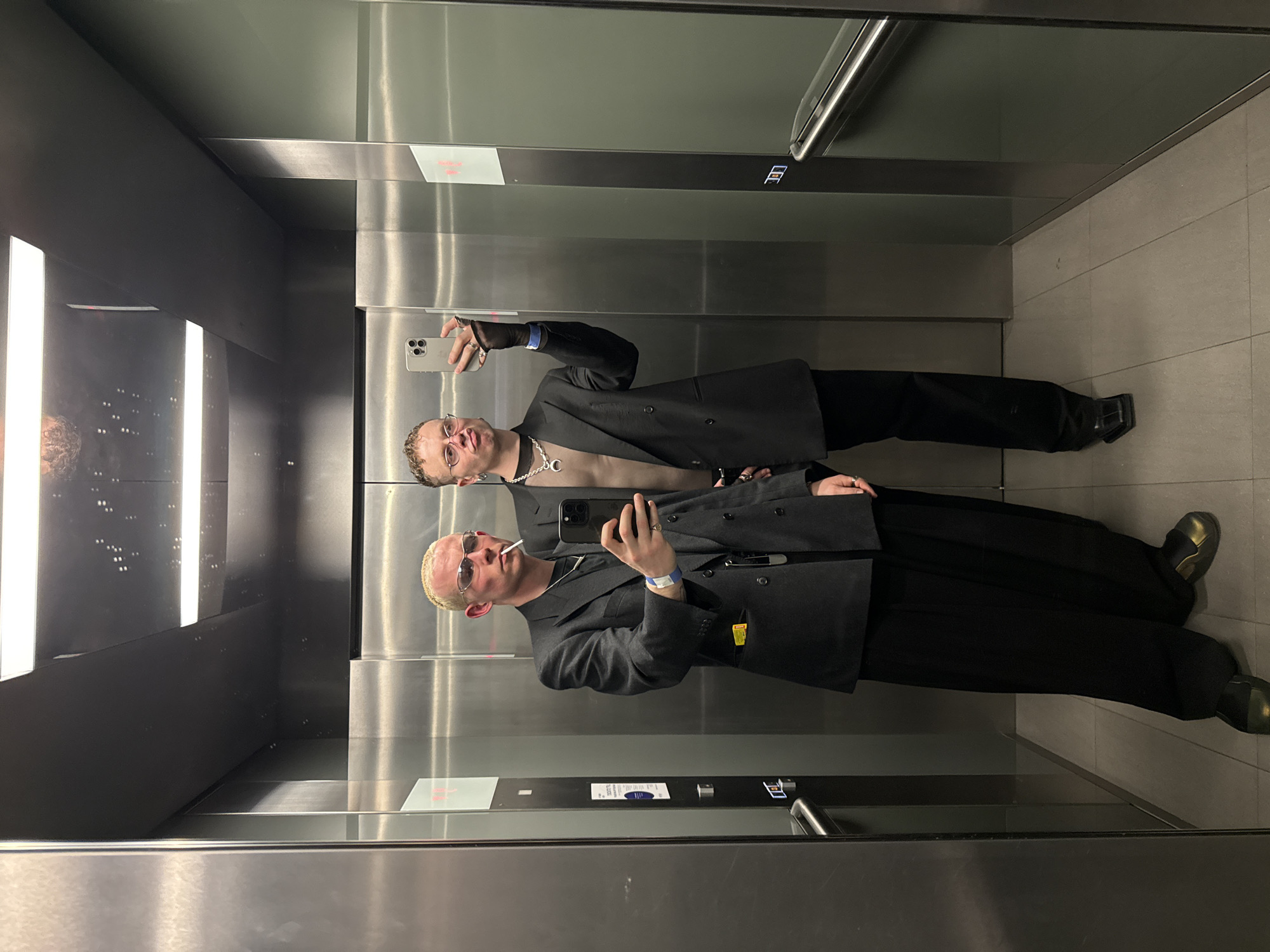 The image, taken in a modern elevator, captures a mirror selfie of two individuals. The setting features brushed metallic walls and a light-colored tiled floor, with vertical strip lighting reflecting in the highly polished surfaces.
Two young men are visible in the reflection. In the foreground, one man with short, light-colored hair is wearing sunglasses and has a cigarette in his mouth. He is dressed in a dark, possibly grey or black, double-breasted blazer over a lighter-toned top, with a prominent silver chain around his neck. A blue wristband is on his left wrist, and he wears rings on his fingers. He is holding a light-colored iPhone with a triple-lens camera system, presumably taking the photo, which is reflected in the elevator wall. A yellow object is tucked into his blazer pocket.
Behind and slightly above him, the reflection of a second man is visible. He also has short, light-colored hair and wears a similar dark blazer over a light top, with a silver chain. He is also holding a light-colored iPhone.
The activity taking place is the two individuals posing for a mirror selfie within the elevator. The overall mood appears casual and stylish, with attention to fashion details.
Notable details include the modern, well-lit urban elevator environment. On one of the metallic panels, a white rectangular sign with faint red text is visible, which appears to include the word "SMOKING" or related text, possibly an alert or prohibition. Other small, illegible labels and icons are also present on the elevator panel, including what looks like a button panel. The location context is London, United Kingdom.