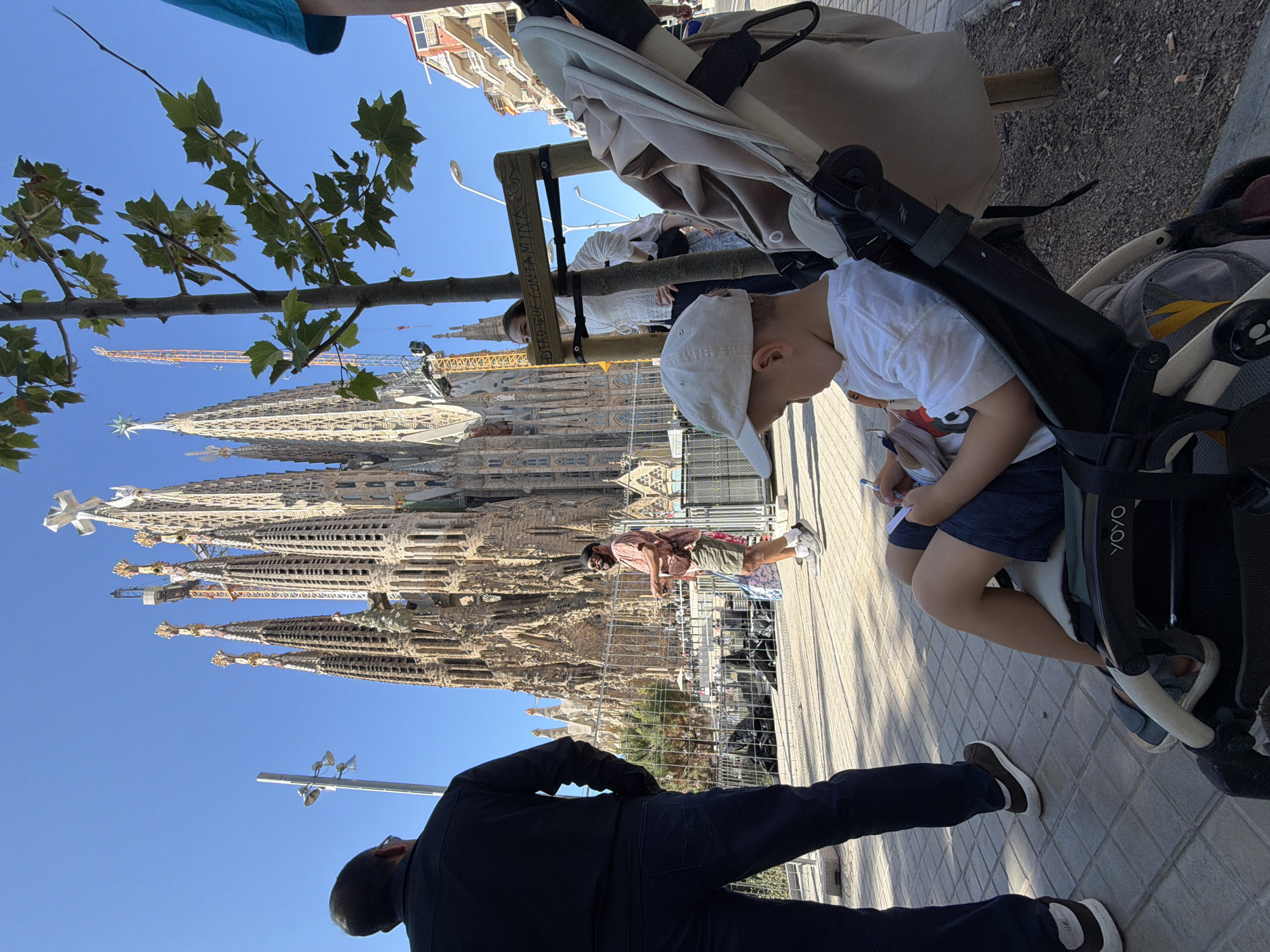 In Barcelona, Spain, in front of the Sagrada Familia basilica, a man in a black shirt and dark pants stands with his back to the camera. To his right, a man wearing a pink patterned short-sleeved shirt, olive shorts, and a fanny pack smiles and holds something in his hand. In the foreground, a young child wearing a white t-shirt and a light-colored baseball cap sits in a stroller, drawing on a piece of paper with a pen. The child's legs are visible, wearing dark shorts. A tree with green leaves is situated between the people and the basilica. A wooden sign is attached to a post near the tree. The sky is clear and blue.