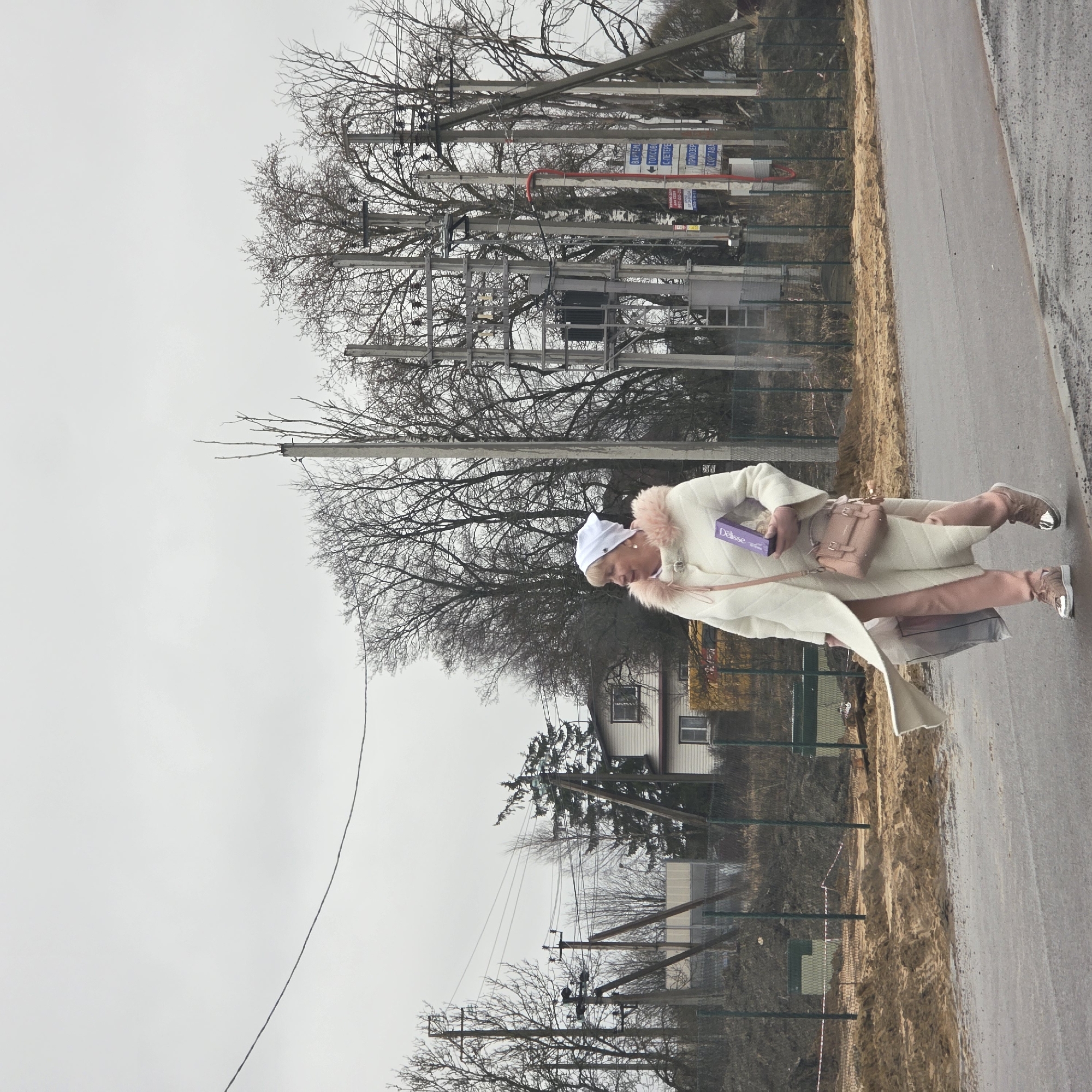The image shows a woman walking along a road in Skotnoe, Russia. She is wearing a white coat with pink fur trim, pink pants, and pink shoes. She is carrying a pink purse and a box labeled "DéLisse". The road is wet and there are bare trees and utility poles in the background. A house and a green fence are visible in the middle ground. The sky is overcast.
