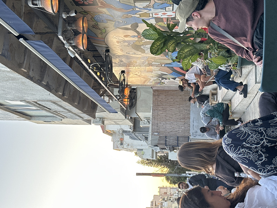 The image captures an outdoor cafe scene at Ecco Cafe in the Florentin neighborhood of Tel-Aviv, Israel, during what appears to be late afternoon or early evening. The setting is a street-level area adjacent to a multi-story concrete building.
Approximately 10-15 individuals, predominantly young adults, are present. In the foreground on the left, a woman with long dark hair, wearing a white top, is partially visible, looking downwards. Next to her, another woman in a dark top and patterned floral pants is seated. On the right, a man in a light green baseball cap and a maroon long-sleeved shirt sits in a dark green outdoor chair, looking to his right.
The mid-ground features several people seated on concrete steps and small wooden benches. Actions include individuals socializing, looking at mobile phones (e.g., a man in a grey hoodie, another in a green hoodie), and one man in a dark shirt smoking a cigarette. In the background left, a man in a dark jacket stands, looking off-camera.
The cafe wall is adorned with a large, colorful mural featuring abstract, stylized human figures and geometric patterns in various hues. Above the mural, a dark blue retractable awning is extended, from which multiple industrial-style pendant lights with warm orange glows and black fan-style heaters are suspended. A large potted plant with broad green leaves is positioned in the mid-ground, to the right. The background shows additional concrete residential buildings with balconies and air conditioning units, along with a bamboo-like privacy screen and a tall conifer tree. The bright, warm light on the left horizon indicates a low sun, possibly sunset.