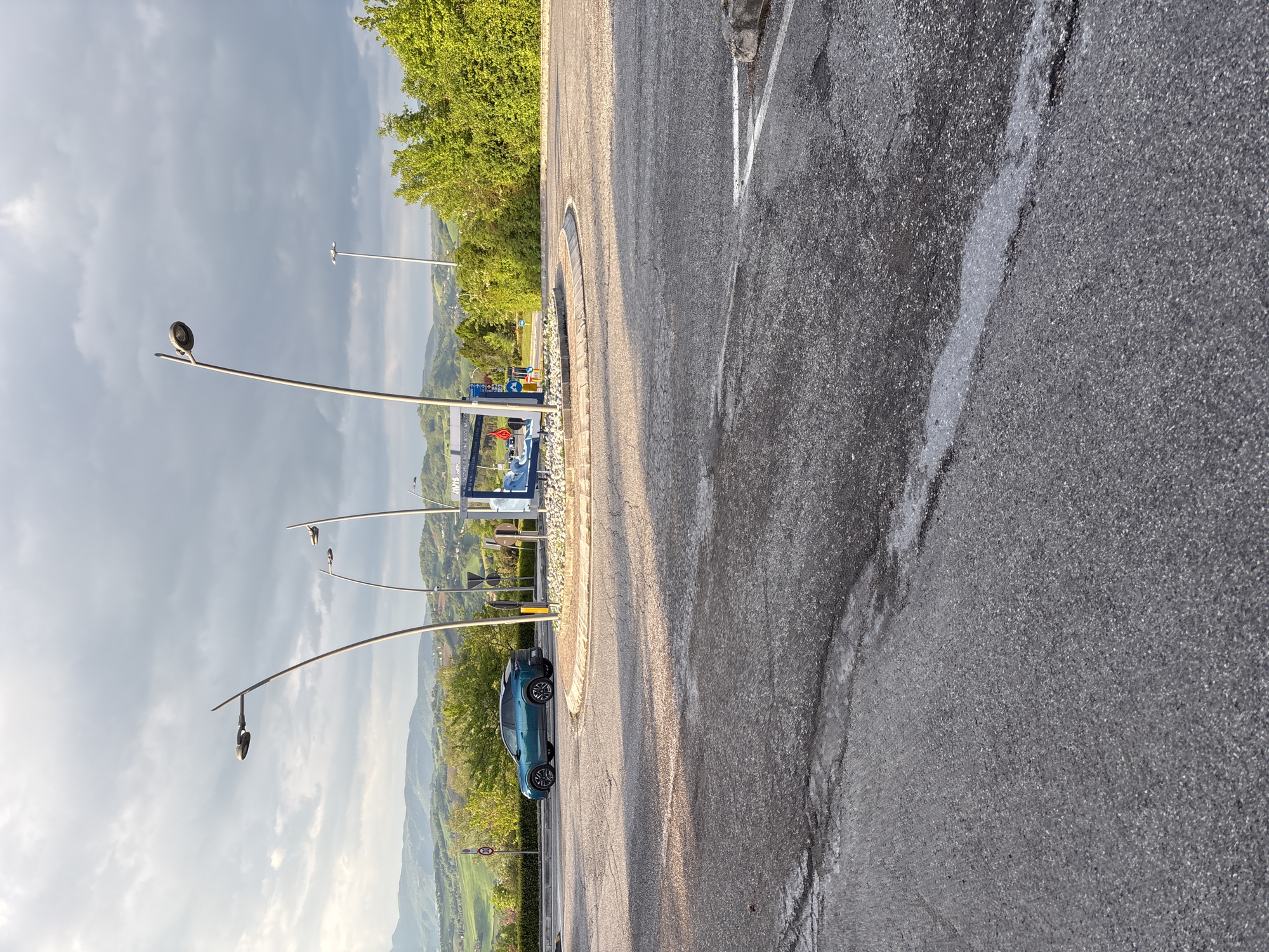 The image captures an outdoor scene, likely a parking lot or a service area, set against a backdrop of rolling hills and lush greenery, characteristic of the Camerino region in Italy. The foreground and midground are dominated by asphalt pavement, textured and dry, with some faint white line markings. Long shadows are cast across the ground, indicating either early morning or late afternoon.
Several modern-style streetlights, featuring slender, curved metallic poles and circular lamp heads, punctuate the left side of the frame. In the midground, a blue car, resembling an SUV or crossover, is parked alongside a curbed island. Dense green trees and vegetation form a natural boundary on the right and in the background, contrasting with the gray asphalt and sky.
The distant landscape reveals multiple layers of green hills and mountains extending towards the horizon under a partially cloudy sky. In the mid-distance, a blue sign with white lettering is partially visible, clearly showing "AVIS," suggesting a car rental service. Below "AVIS," another segment of text, possibly "STAT" or "SERVIZI," is faintly discernible. There is no visible human activity, lending a peaceful and static quality to the scene. A small, round traffic sign is also present near the blue car, though its specific details are unreadable.