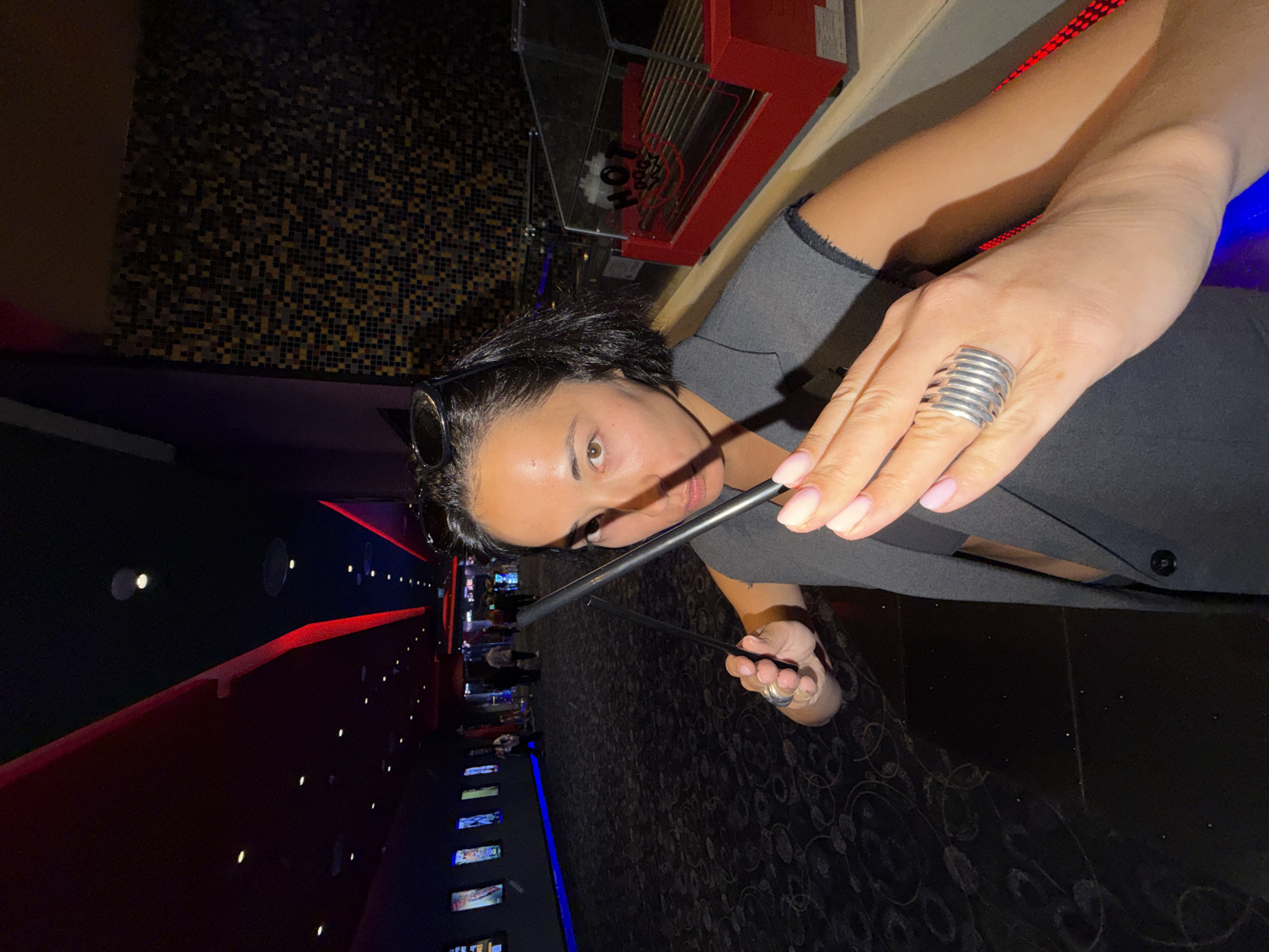 The image shows a close-up of a woman in a movie theater. She is holding two black straws and looking towards the camera. She has dark hair, brown eyes, and is wearing a gray top and a large silver ring on her right hand. The background is dark, with the red and blue lights typical of a cinema hallway visible, along with the patterned carpet and the wall of a popcorn machine. The scene appears to be indoors, likely at night, given the dim lighting of the theater. The woman's expression is neutral. The location is identified as Rishon LeZion, Israel.