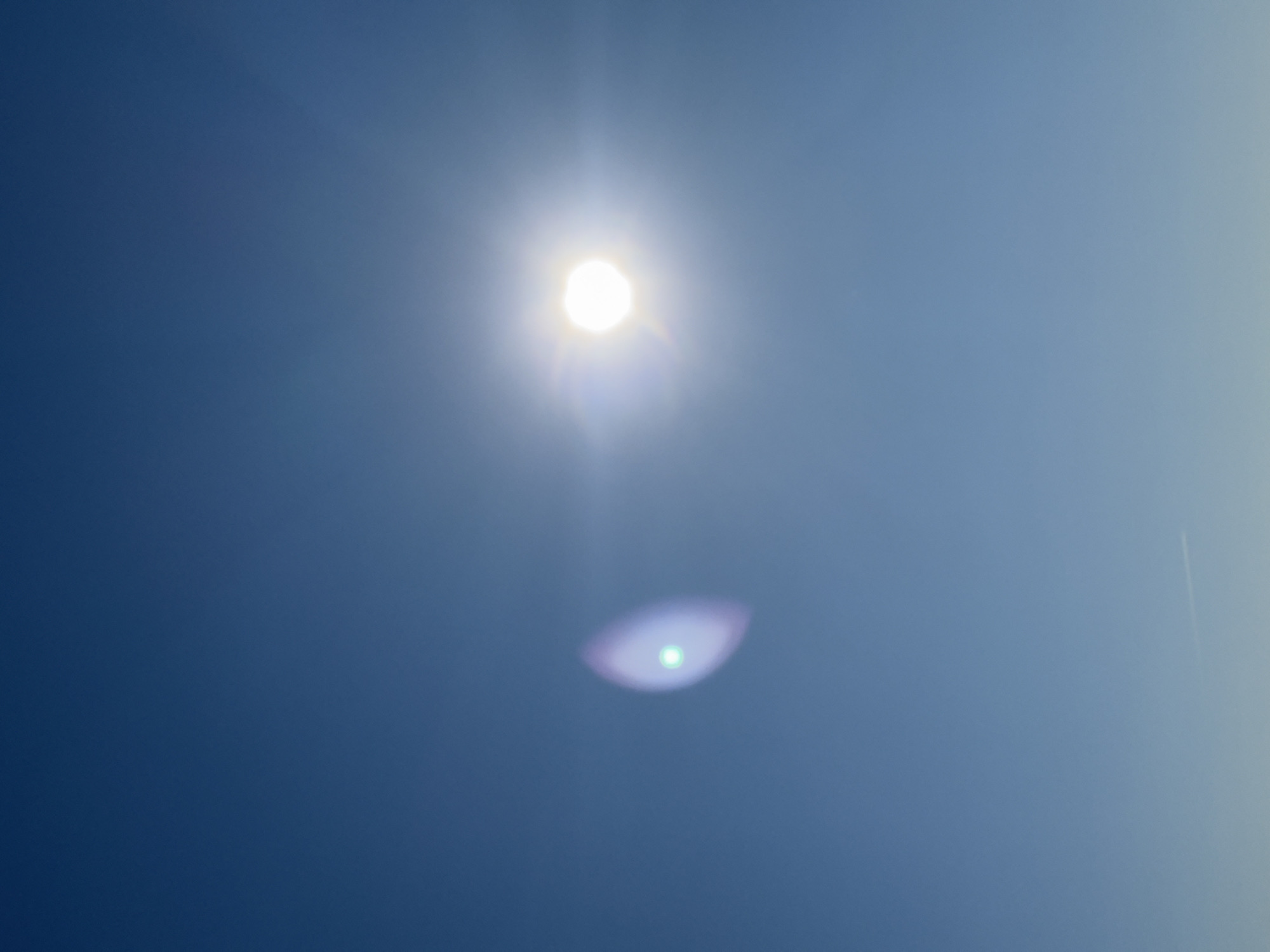 The image shows a view of a clear blue sky with the sun prominently visible at the center. A lens flare is present below the sun. The image does not provide any location-specific details that would confirm it was taken in Votkinsk, Russia. There are no people, subjects, objects, actions, or interactions identifiable in the image, other than the presence of the sun in the sky. The environment is simply a clear, cloudless, blue sky.