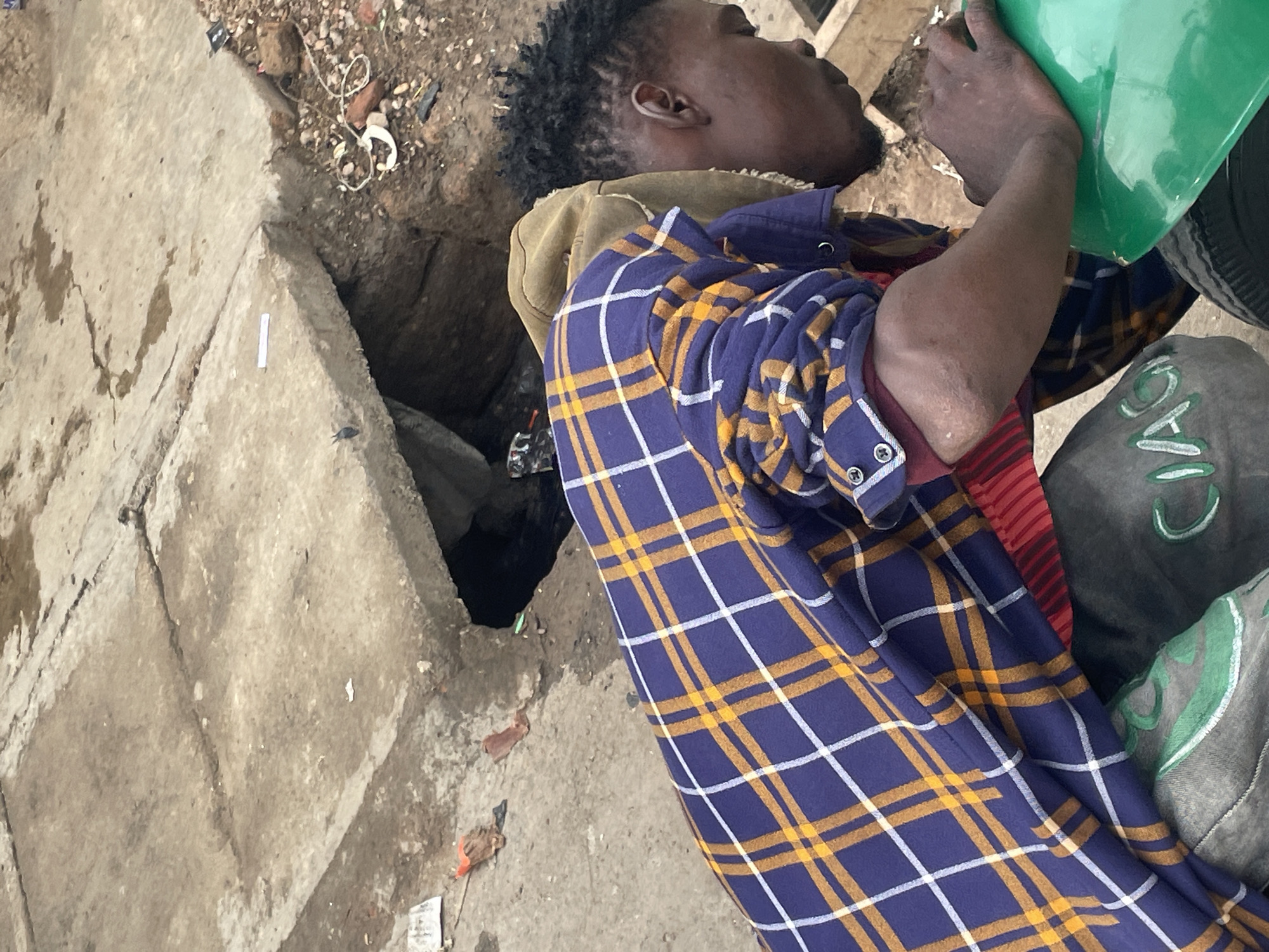 A man is drinking from a green container while positioned near a hole in the ground. The man has dark skin, dreadlocks, and is wearing a blue and yellow plaid shirt over a red striped garment and a tan hooded sweatshirt. He appears to be on the ground near a concrete surface with debris and trash. The hole in the ground suggests a possible drainage or sewage system. There is additional trash scattered on the ground nearby. The image was taken in Jos, Nigeria.