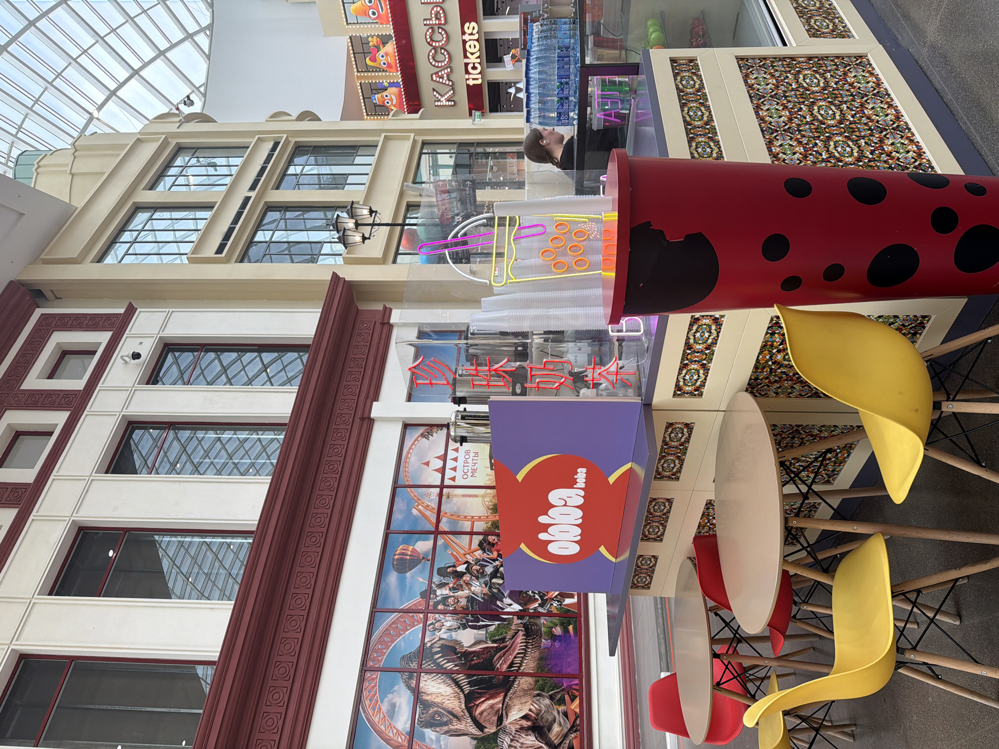 This is an outdoor shot taken in what appears to be a shopping mall or entertainment complex, under a large, glass-domed ceiling. The focus is on a boba tea kiosk with a vibrant, red trash can decorated with black circles, resembling a ladybug, positioned in the foreground. The kiosk itself has a purple panel with the "obba boba" logo. Behind the kiosk, a large mural depicts a dinosaur and a roller coaster. Several round tables with red and yellow chairs are set up in front of the kiosk. In the background, a store with "tickets" signage and cartoon characters is visible, along with rows of bottled water and a woman working behind the counter at the kiosk. The architecture features ornate columns and large windows reflecting the glass ceiling.