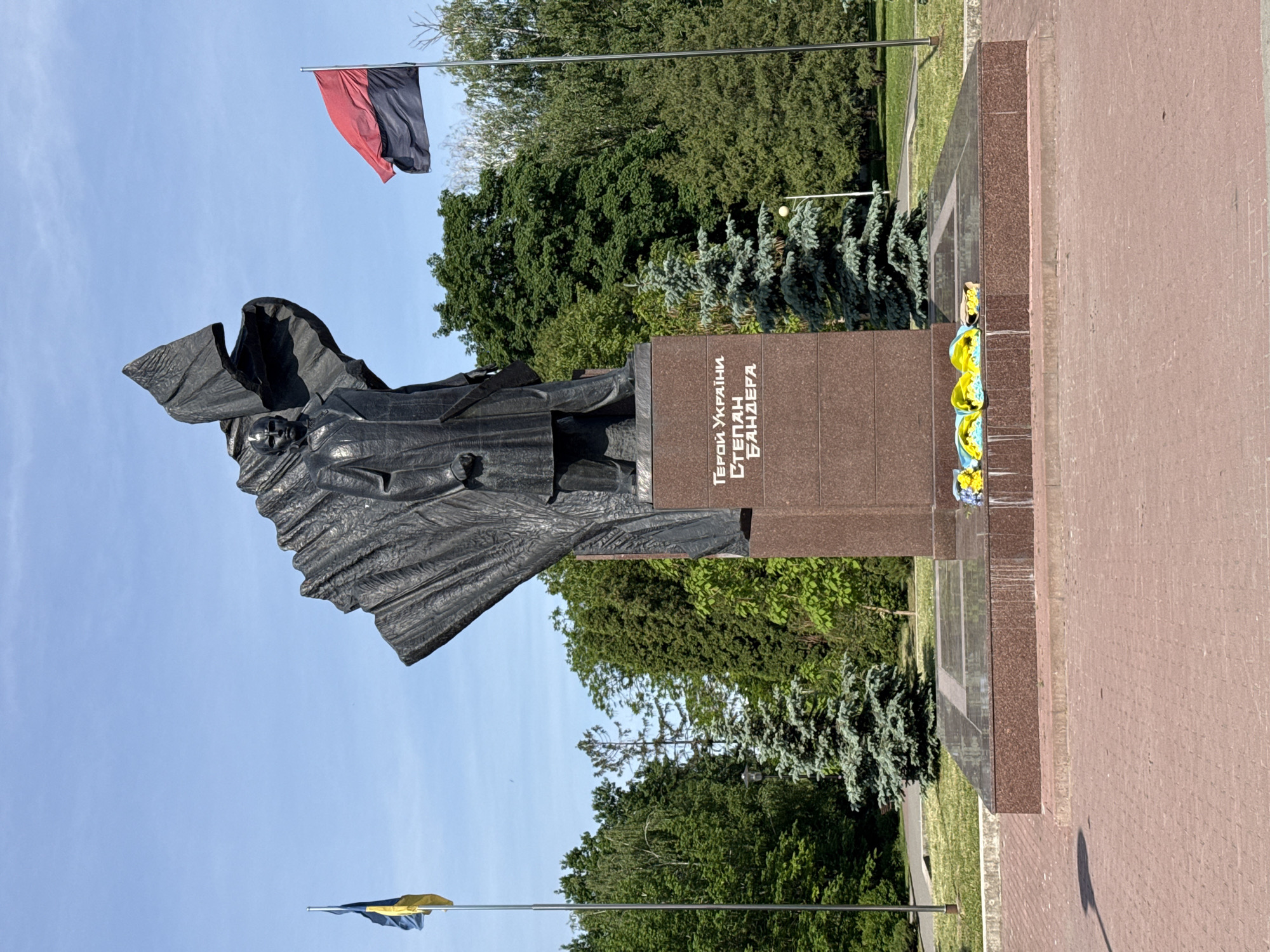 A statue of Stepan Bandera, a Ukrainian political activist and theorist, stands in the city of Ternopil, Ukraine. The statue is made of dark metal and shows Bandera with a determined expression. The statue is mounted on a brown marble pedestal, which is decorated with blue and yellow flowers, the colors of the Ukrainian flag. Two flags are flying in the background. One is black and red, the flag of the Ukrainian Insurgent Army (UPA), and the other is blue and yellow, the flag of Ukraine. The statue is located in a park, and trees are visible in the background. The sky is blue.