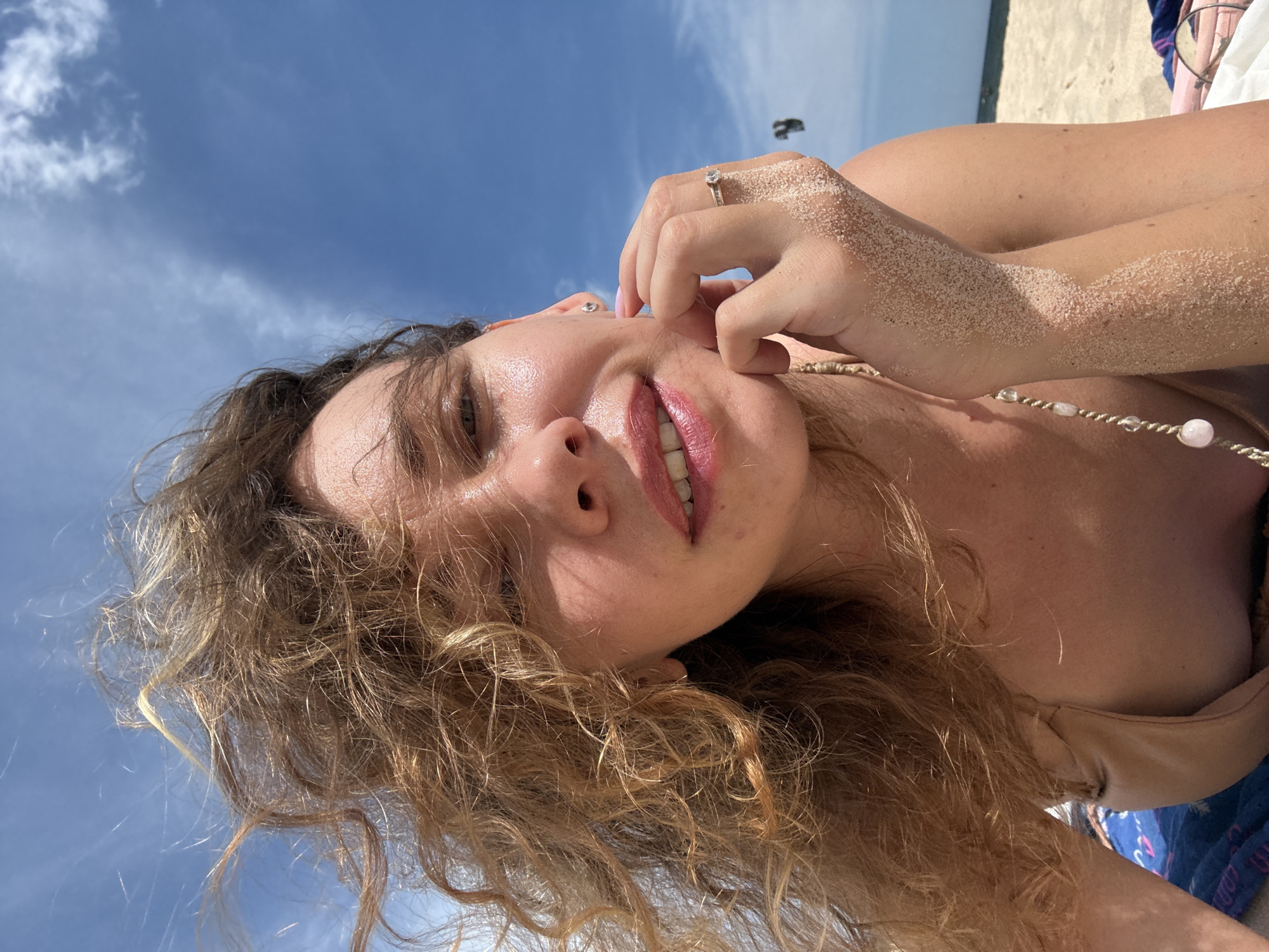 An outdoor, close-up, low-angle selfie of a woman with medium-length, curly, light brown hair taken on a beach with a bright blue sky and scattered white clouds. The woman is looking towards the camera with a slight smile and has a tan complexion. She is wearing a tan bikini top and has sand on her right hand and arm. A ring is visible on her right ring finger. The background shows a sandy beach and the ocean. A dark object, possibly a kiteboarder, is visible in the distance. The lighting suggests it is daytime with strong sunlight.