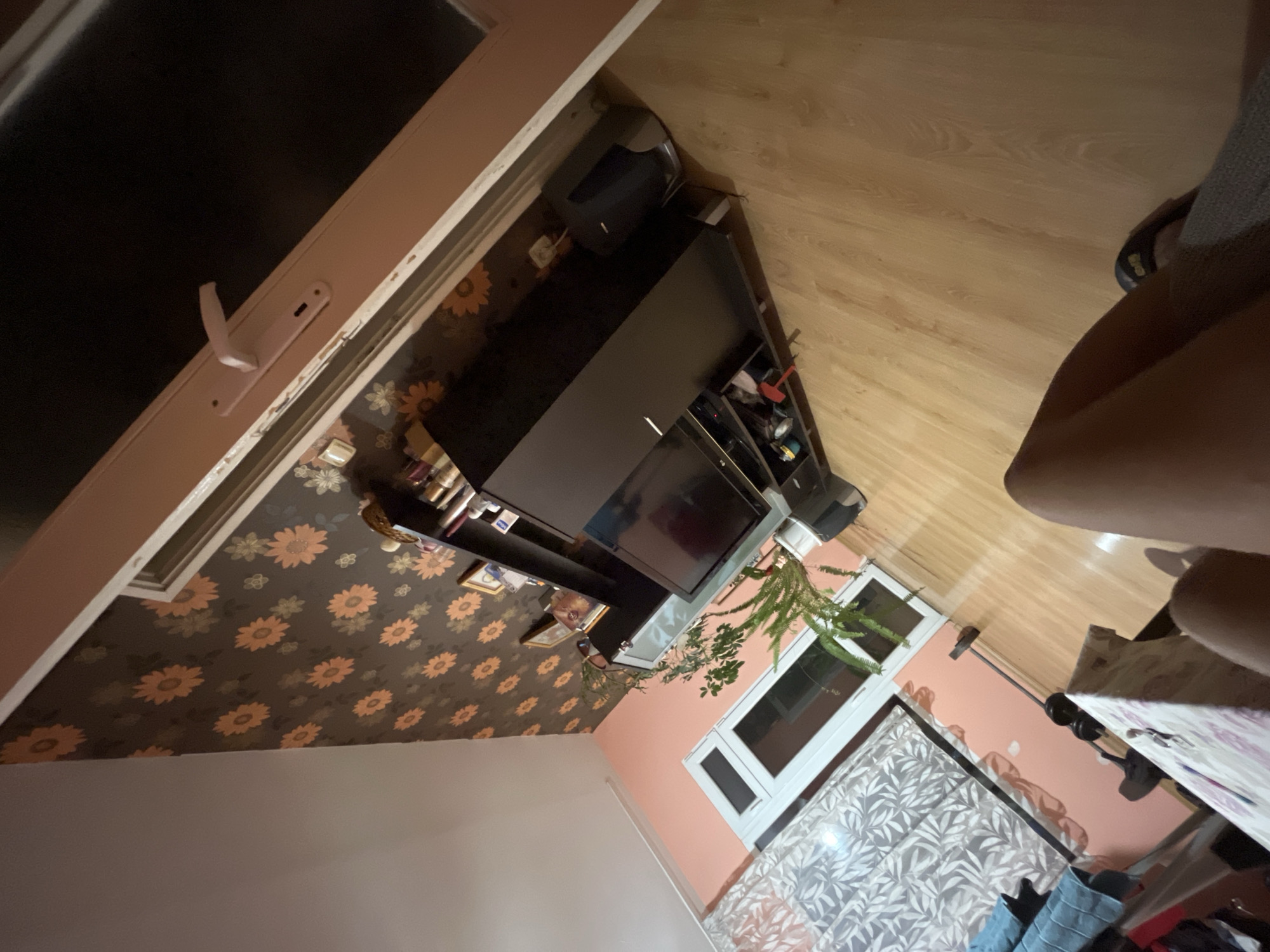 The image depicts an indoor scene, likely a living room, with a focus on a media console and a table. The room features light brown wooden flooring and walls painted in a soft peach color, with one accent wall covered in wallpaper featuring a repeating pattern of orange flowers against a dark brown background. A large window with white trim and patterned curtains is visible on the left.
On the floor, a black media console stands against the floral wallpaper. It supports a dark flat-screen television. Above the television, shelves are filled with various items, including framed pictures, bottles, and decorative objects. A potted green plant with fern-like leaves sits in a silver pot next to the media console, positioned on a stand.
To the left of the media console, a white table with a patterned tablecloth is partially visible. A person's legs and feet, wearing dark sandals, are in the foreground, suggesting they are seated or standing near the table. A barbell with weights is visible on the floor behind the table, hinting at exercise equipment.
The lighting appears to be artificial and somewhat dim, casting shadows and creating a cozy atmosphere. The overall impression is of a lived-in space. The specific location of Plovdiv, Bulgaria, is not discernible from the visual elements in the image itself.