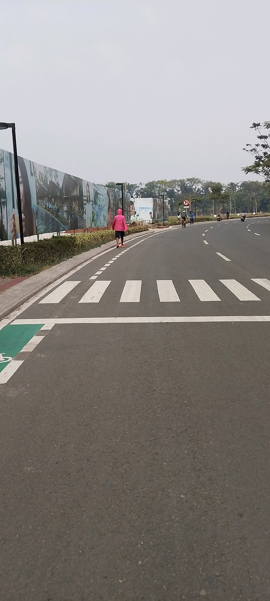 In Gunung Putri, Indonesia, a person in a bright pink jacket walks along a paved road. The road features a clearly marked pedestrian crossing with white stripes. To the left of the road, a large mural or banner stretches along a wall, depicting various scenes. Adjacent to the mural is a strip of green vegetation. Further down the road, other individuals can be seen, some on bicycles and others on motorcycles. The environment appears to be an outdoor urban or park area with trees visible in the background and a cloudy sky overhead.