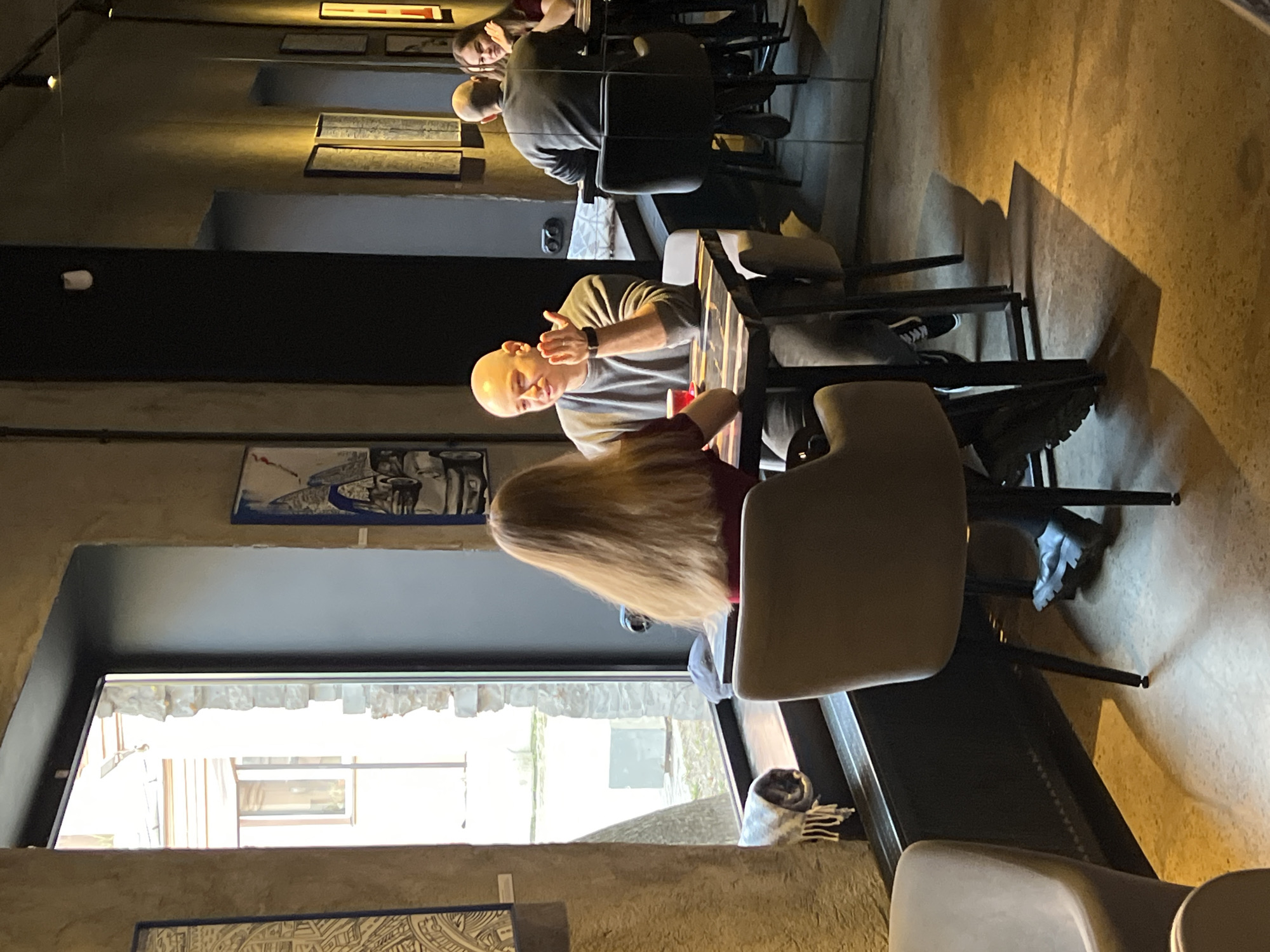 Here is a detailed description of the image:
**Scene:** Indoor setting, likely a restaurant or cafe.
**People:** A bald man in a grey shirt is seated at a table, gesturing with his hand. A woman with long blonde hair is seated across from him. Additional individuals are visible in the background, seated at other tables. Reflections in the wall mirrors show more people.
**Objects:** Tables, chairs, plates, and possibly other dining-related items are visible. Additionally, artwork is on the walls, and there is a window letting in natural light.
**Actions:** The bald man appears to be speaking or explaining something. The woman is seated and likely listening. Other people in the background are seated and seemingly engaged in conversation.
**Interactions:** Appears to be a conversation between the man and the woman.
**Environment:** The interior is designed with a modern aesthetic, featuring artwork and mirrors on the walls. The window provides a view of buildings outside.
There is not enough information to specify the location to the city of Saint Petersburg, Russia.