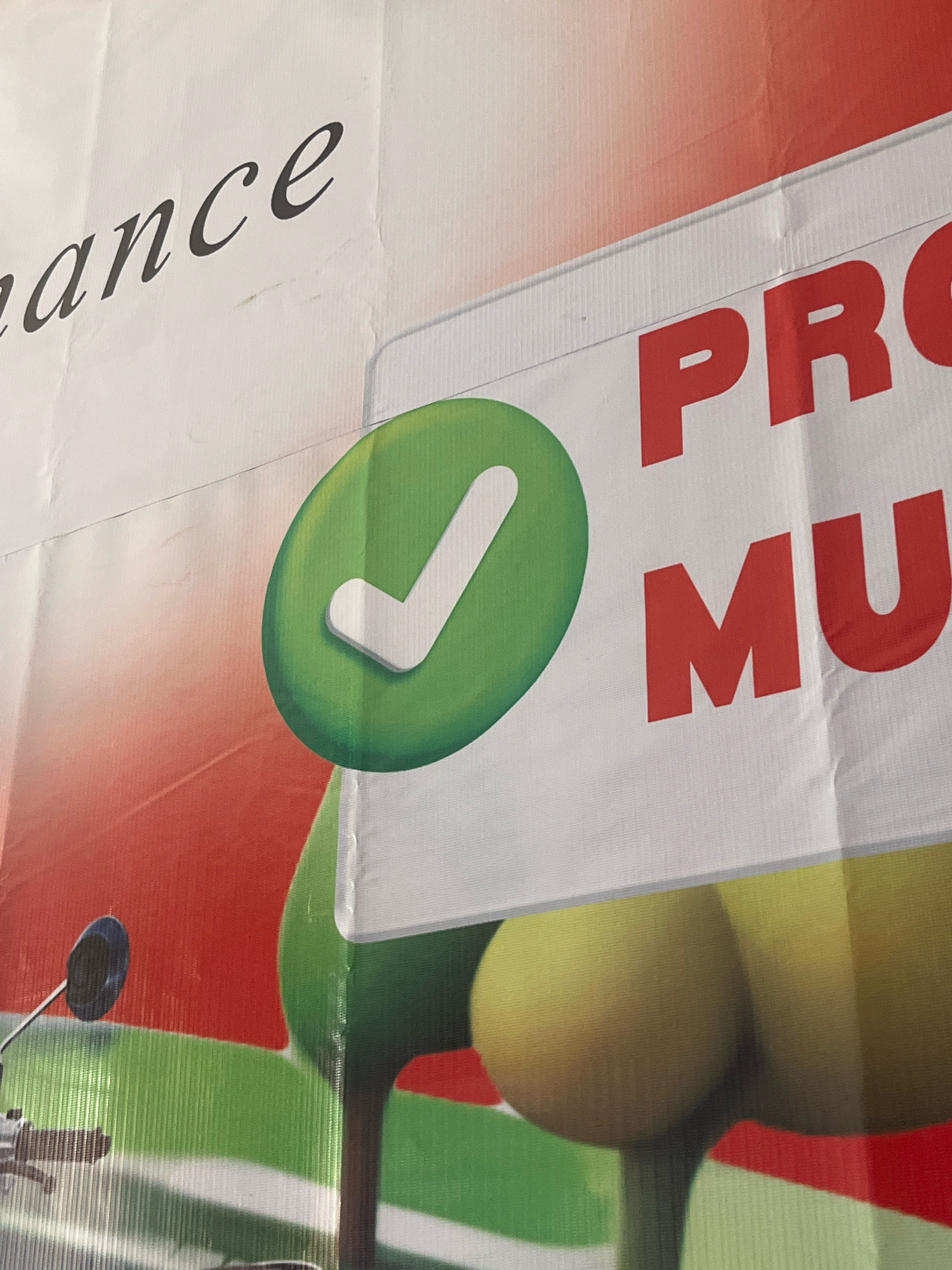 A close-up, eye-level shot captures a portion of a banner with bold red lettering. The banner is predominantly white, with a gradient of red and orange filling the upper portion of the image, fading into a red hue as it ascends. To the left, a large, vibrant green checkmark is encased within a circular green border. The checkmark is outlined in white, giving it a three-dimensional appearance.
The red text on the banner is large and blocky. The letters "PR" are clearly visible on the upper line, and "MU" are visible on the lower line. The letters appear to be at least one foot tall. The banner is creased and wrinkled in several places, suggesting it is made of a flexible material like vinyl or fabric.
In the lower left quadrant of the image, a black rearview mirror with a metallic arm is visible, attached to a handle that is partially obscured. The background behind the mirror is a vivid red, and below that, a striped green and white pattern representing grass. Further to the right, two large, round, yellow objects, possibly stylized fruit like apples or lemons, are depicted. The banner is likely displayed outdoors, possibly at an event or as part of an advertisement in the city of Boja, Indonesia, though the specific location within Boja cannot be determined from the image alone.