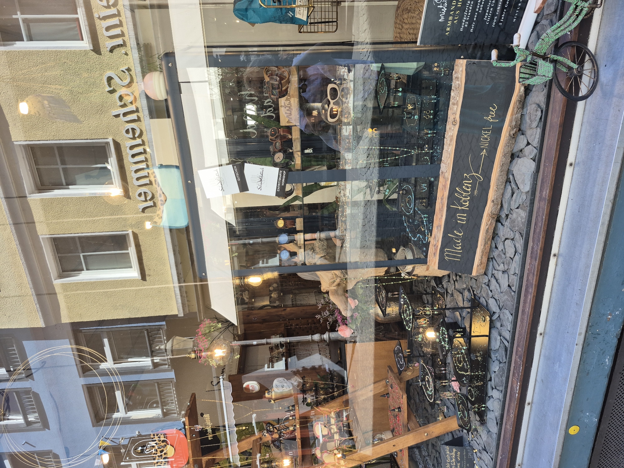 The image shows a storefront window display for a shop called "Schmuck & Stein" in Koblenz, Germany. The window features various jewelry items, including necklaces, earrings, and bracelets, displayed on black stands and a wooden table. Behind the jewelry, there are watches and sunglasses. A sign with the text "Made in Koblenz" and "NICKEL free" is prominently displayed on a rustic wooden board. The shop's name, "Schmuck & Stein", is visible on a sign in the upper part of the window, as well as "MISTURA" and "ARM BÄNDER AUS HOLZ". Reflections of the street, buildings, and the sky are visible on the glass. A small decorative bicycle is placed in front of the display. The overall scene suggests a boutique selling handmade jewelry and accessories.