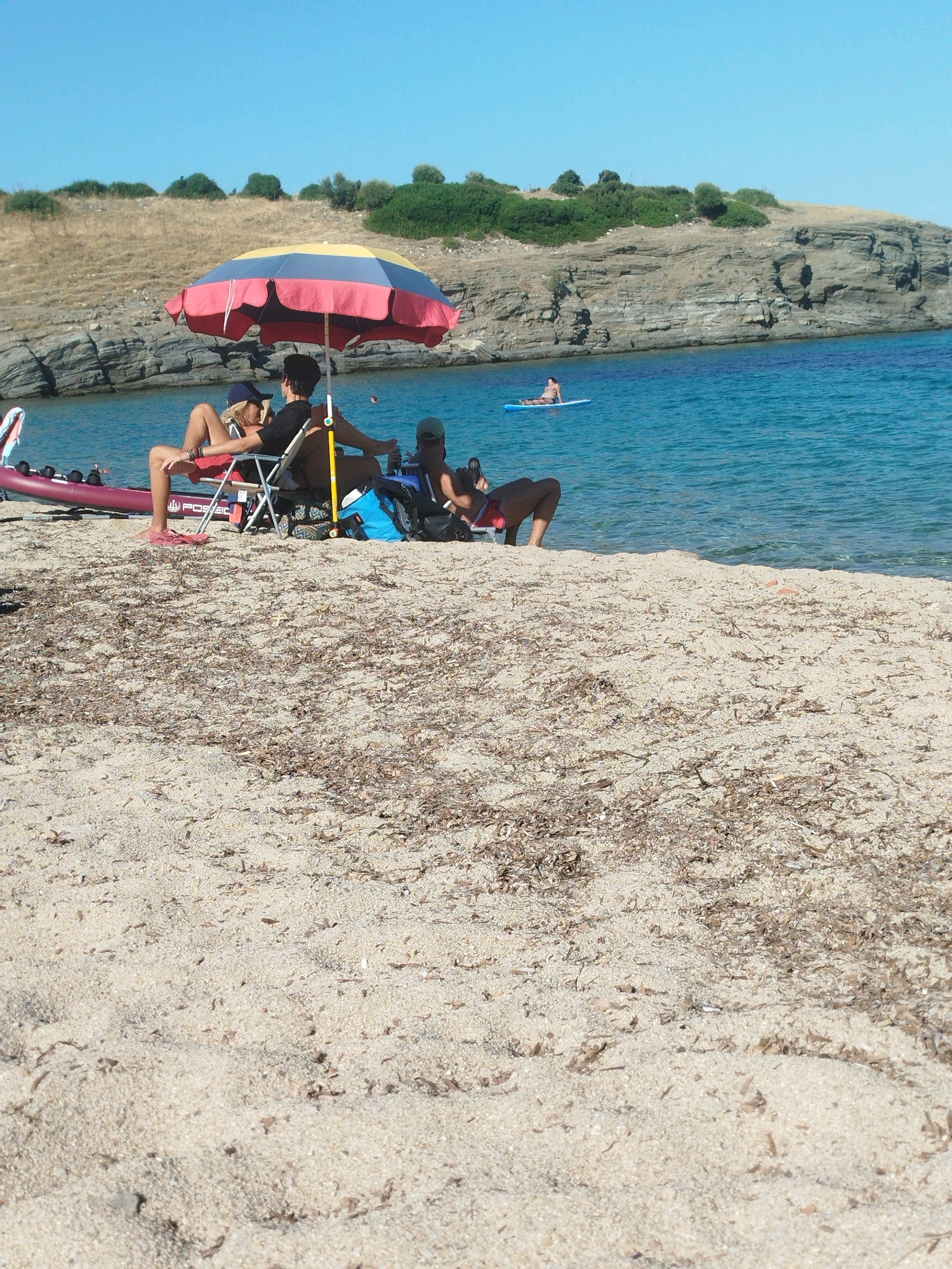 A bright, sunny day at a sandy beach in Sithonia Municipal Unit, Greece, shows several people enjoying the water and shore. In the foreground, the sand is covered with dried seaweed. A colorful beach umbrella, red, yellow, and blue, shelters two people sitting in beach chairs. To the left of the umbrella, a person wearing a blue cap and a bathing suit is leaning back in a red chair, with their legs extended. Next to them, a person in a black shirt and dark shorts is also seated. To the right, another person in a black shirt and dark shorts sits in a beach chair. In the background, the clear blue water of the sea is visible. Further out in the water, a person is lying on a paddleboard. Behind the water, a rocky, vegetated hillside slopes up towards a clear blue sky.