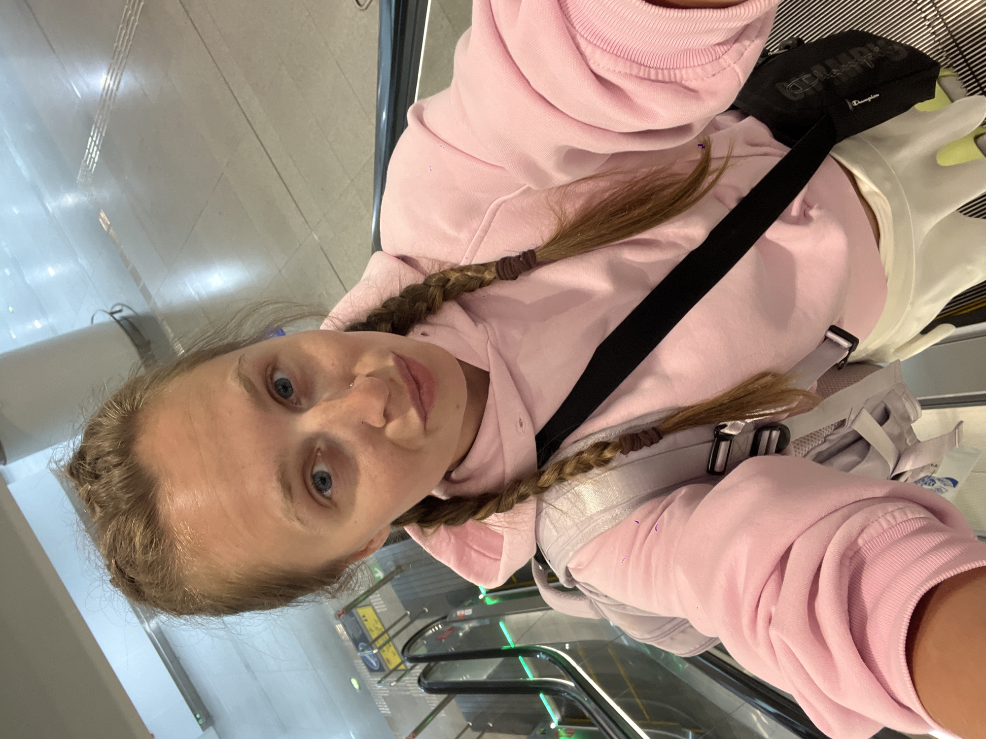 A young woman with blonde hair styled in two braids stands on an escalator. She is wearing a light pink hooded sweatshirt and a white skirt. A black crossbody bag is slung over her shoulder, and she has a light grey backpack on. The background shows the interior of what appears to be a transportation hub, with a pillar and tiled flooring. The escalator's steps and handrails are visible. The image is a selfie, with the woman looking towards the camera. The scene is located in the city of Nong Prue Subdistrict, Thailand.