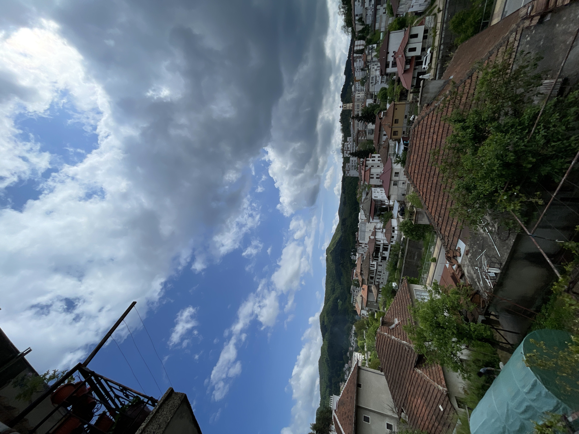 The image shows an elevated view of Zlatograd, Bulgaria. The foreground features residential buildings with terracotta roofs and verdant trees. A mountain ridge densely covered with trees runs vertically through the frame. Beyond the ridge, a dense cluster of buildings spans the skyline. The sky transitions from deep blue with scattered cumulus clouds to dark gray storm clouds. A metal structure, possibly a balcony, appears in the lower left corner.