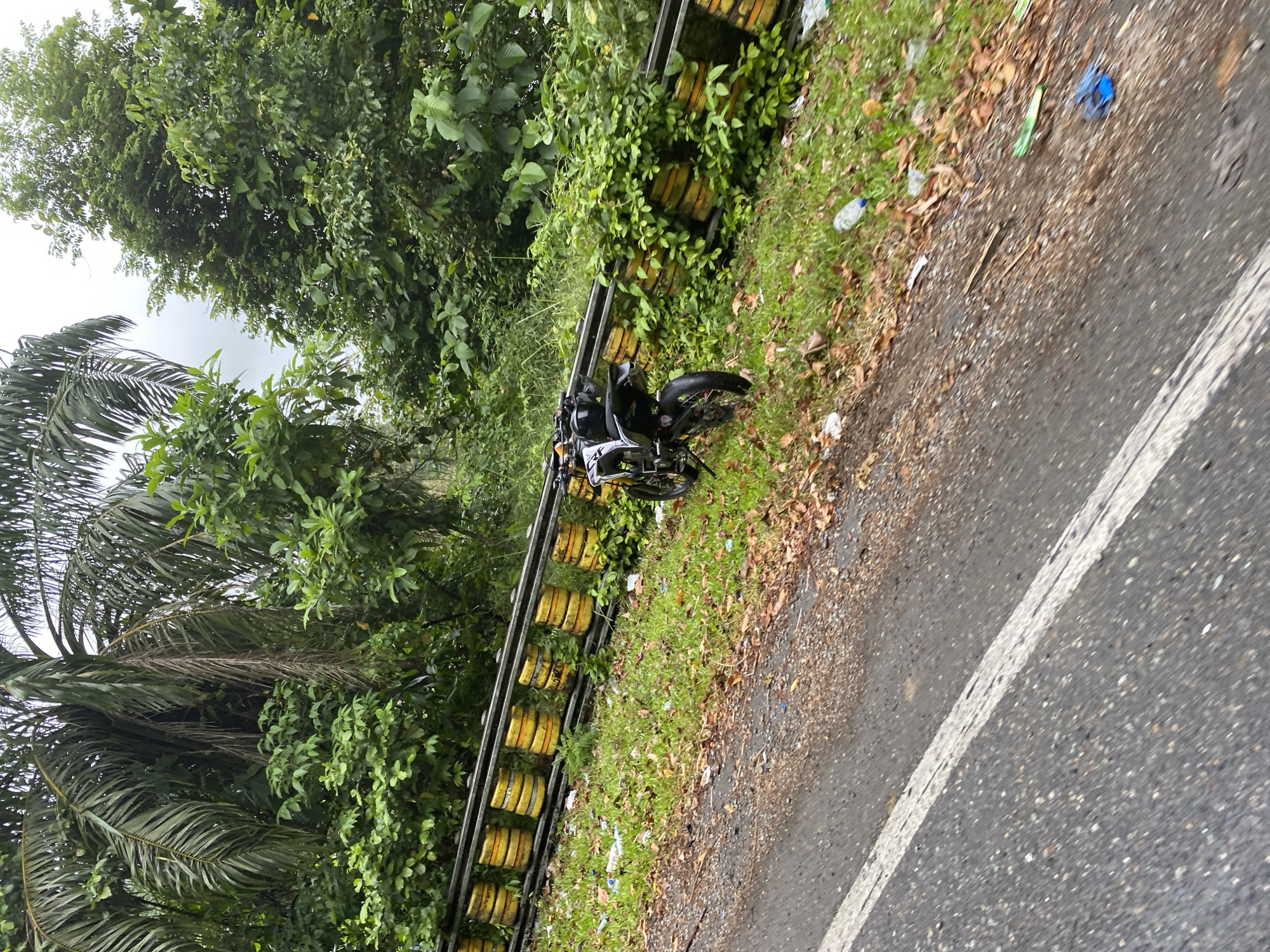 A motorcycle is parked on the grassy shoulder of a road, next to a yellow and black barrier. The barrier appears to be made of stacked cylindrical objects. Lush green trees and palm fronds are visible behind the barrier and extend into the background. The road surface is asphalt with a white line marking its edge. Scattered on the grass are fallen leaves and plastic debris, including bottles and wrappers. The sky is overcast. The location is the city of Babahie, Indonesia.