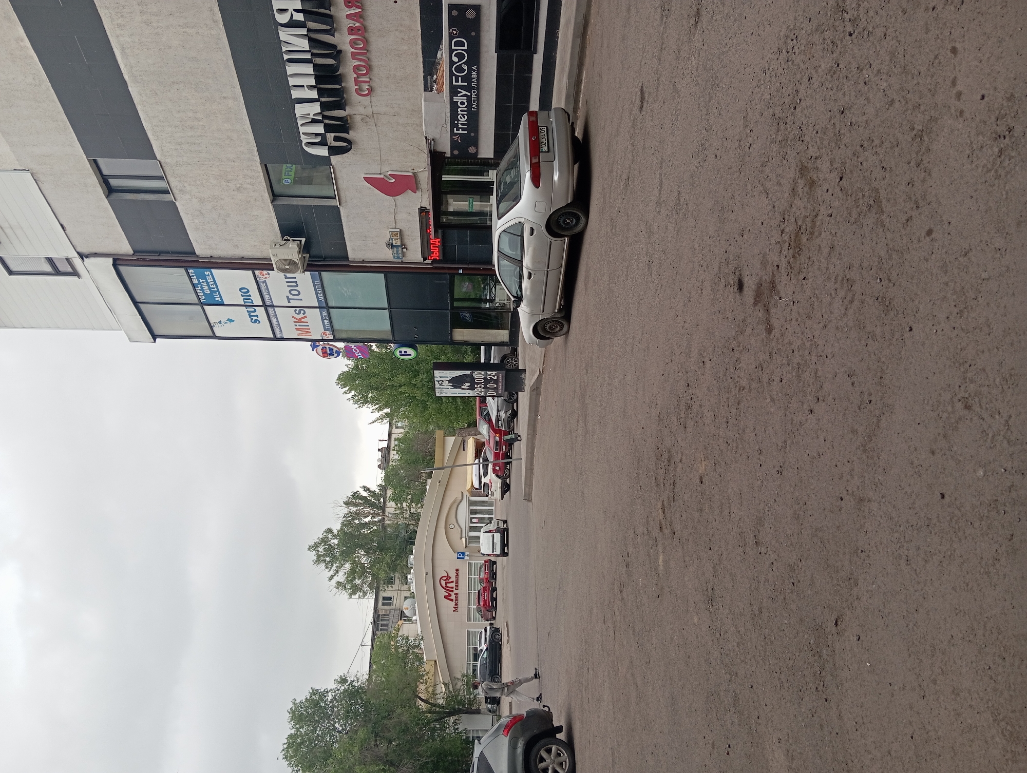 The image captures a street scene in Karaganda, Kazakhstan, showing a building with commercial signage and parked cars. The building features signs for "СТАНЦИЯ СТОЛОВАЯ" (Station Canteen) and "Friendly FOOD" (Gastropub). Below, there is a sign listing "TOEFL, IELTS, GMAT ALL LEVELS" and "Miks Tour," a travel agency. A silver car with license plate "102LNA 09" is parked along the curb. Further down the street, another building is visible with the sign "Мясной павильон" (Meat Pavilion). Various cars are parked along the street. The sky is overcast.