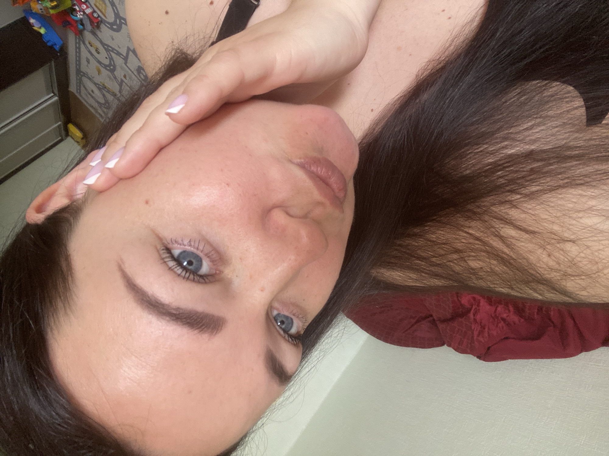 A close-up, rotated image shows a woman with fair skin, dark hair, and blue eyes. She is partially obscured by her own hand. Her fingernails are manicured with a French tip. The background includes what appears to be a drawer and a patterned surface with toys. A red fabric is visible below her hair. The image provides no information that indicates it was taken in Sochi, Russia.