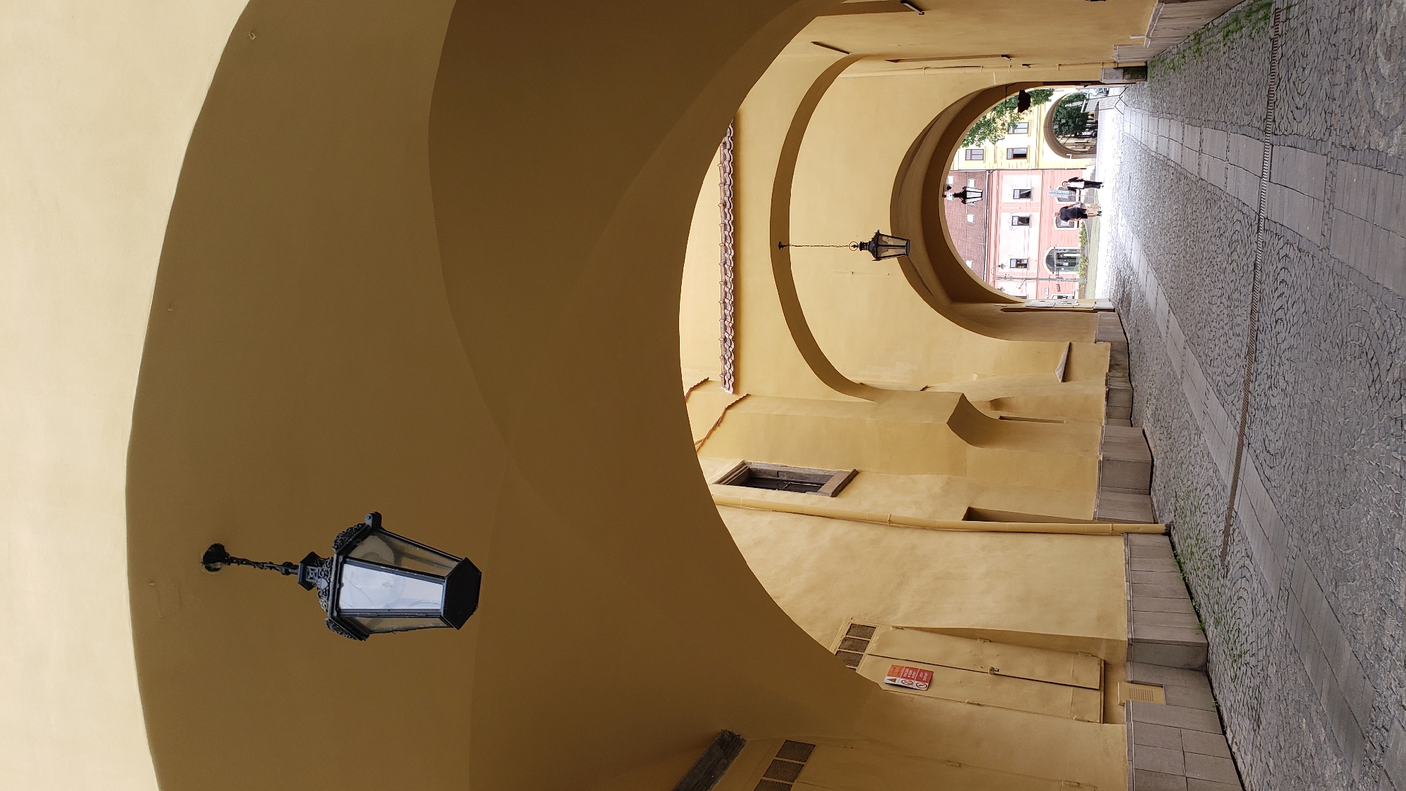 The image depicts a view through a series of arched passageways, characteristic of the historical architecture found in Prešov, Slovakia. The passageways are constructed from yellow-hued stone or stucco and feature vaulted ceilings. Two ornate, black metal lanterns with glass panes are suspended from the ceilings within these arches. The ground is paved with cobblestones and interspersed with lighter stone pathways.
In the background, the passageway opens onto a square or street lined with buildings. One building in the mid-ground is a multi-story structure with a red-tiled roof and several windows. Further in the distance, additional buildings are visible, suggesting an urban setting. Two individuals are walking away from the camera, towards the open area in the background, one appearing to be a man in a dark shirt and shorts, and the other a woman in a dark outfit. The overall lighting suggests it is daytime, possibly overcast or late afternoon given the soft shadows. The scene conveys a sense of historical depth and urban charm.