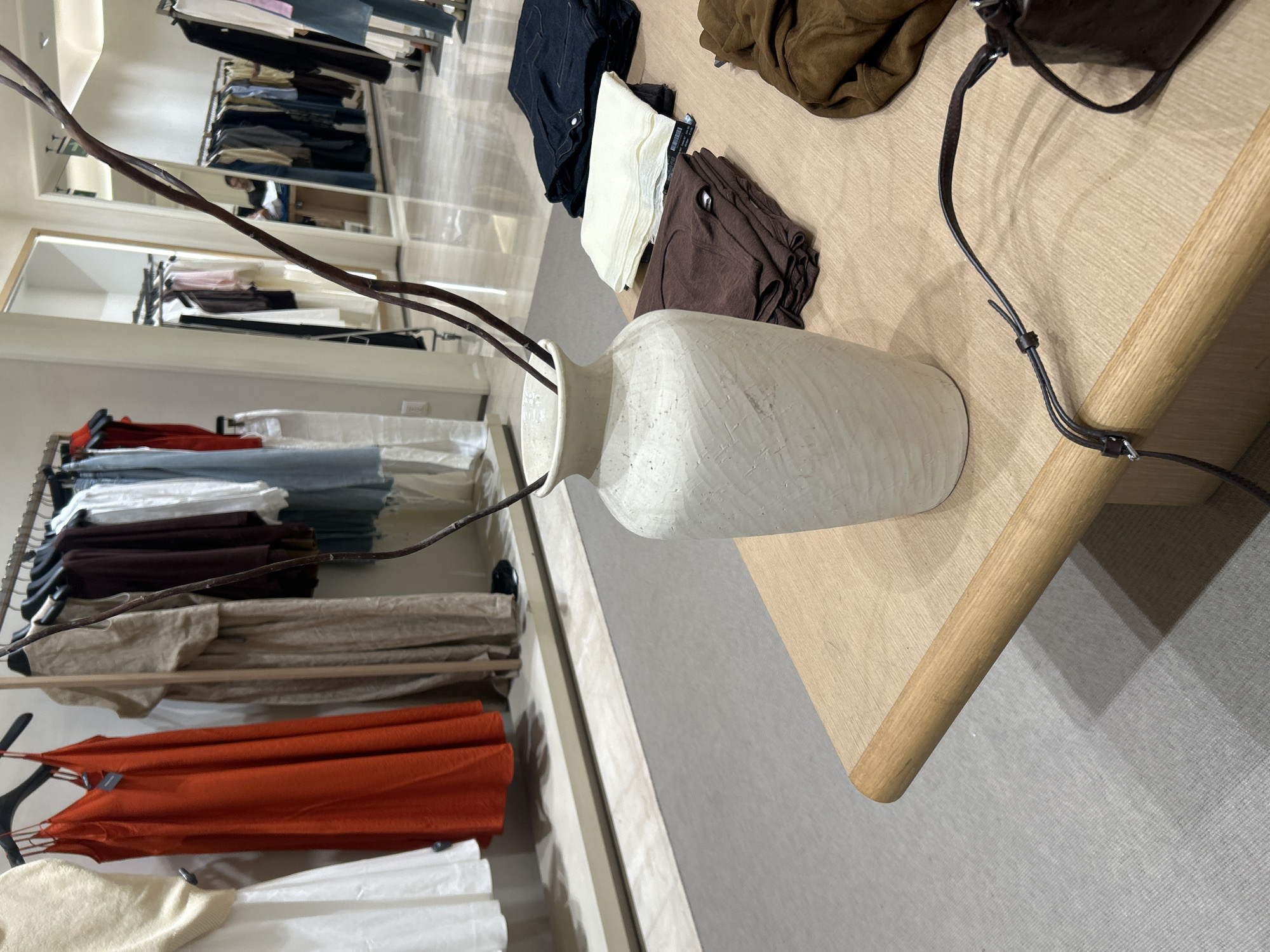 The image shows an indoor clothing store setting, location unknown. A white, textured vase with dried branches sits on a light wooden display table. On the table are folded clothing items in shades of black, white, and brown. A dark brown leather purse with a strap also rests on the table. In the background, clothing racks display items in various colors, including red, orange, white, and shades of blue. The floor is a neutral gray. The overall impression is a curated display in a retail environment.