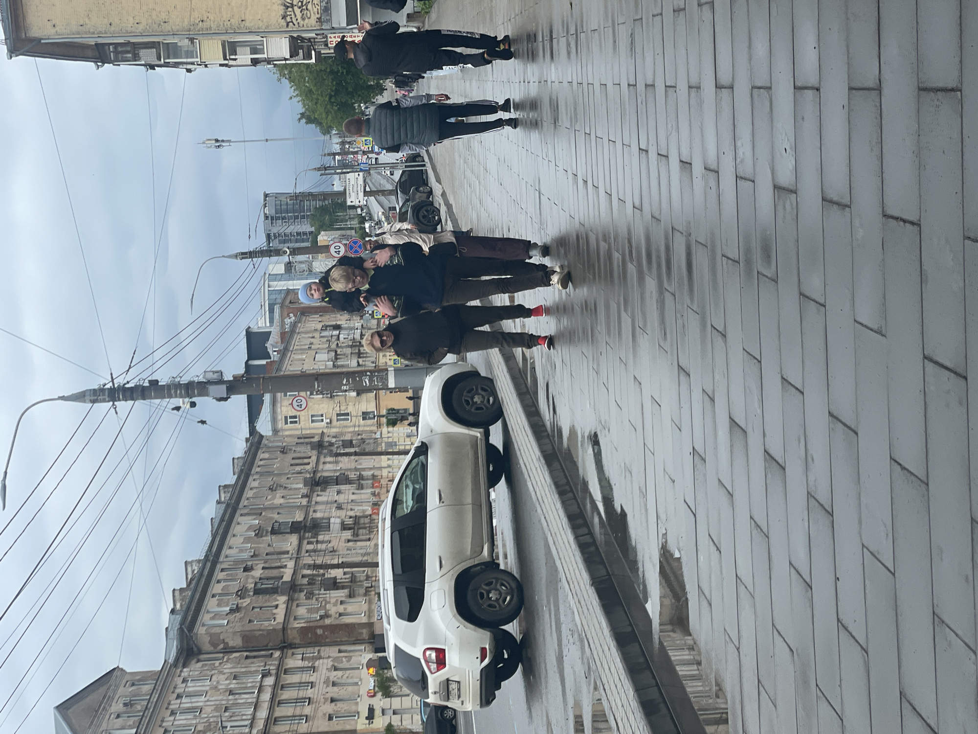 The image depicts a street scene in Izhevsk, Russia. The photograph is taken at street level. A group of people are walking along the sidewalk, appearing to be together. A white car is parked along the side of the road. The street is paved with rectangular gray stones. In the background, there are several buildings, including an older building with multiple windows and a newer, taller building. The sky is overcast, suggesting a cloudy day. A street lamp and power lines are also visible. A speed limit sign of 40 is visible in the background. A street sign with text written in a foreign language, potentially Russian, is present.